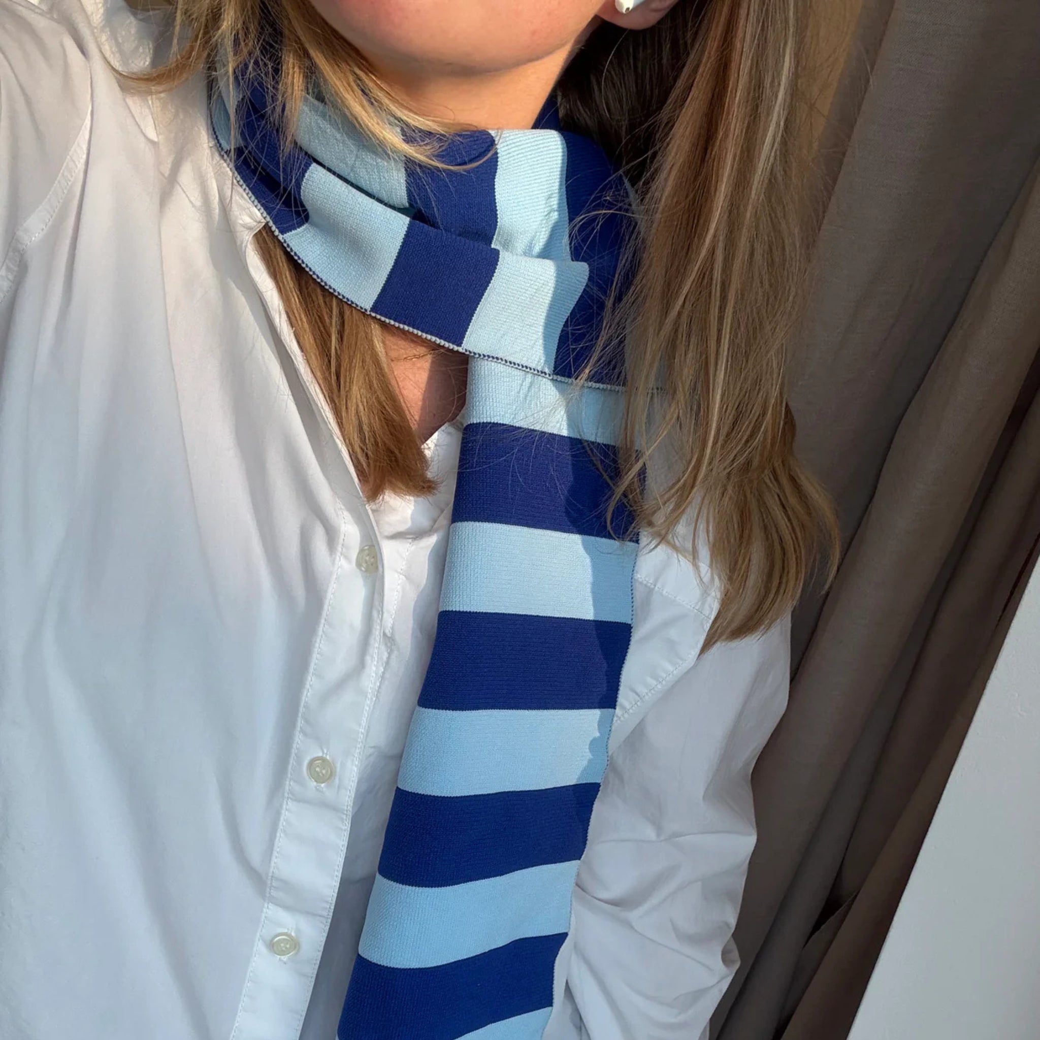 Een persoon in een wit overhemd en de Caro Antwerp Caro Sjaal Ace Ice Ice Baby staat bij een raam, zonlicht op zijn gezicht en schouder. Hun blonde haar zit los en een deel van een bruin gordijn is zichtbaar op de achtergrond.