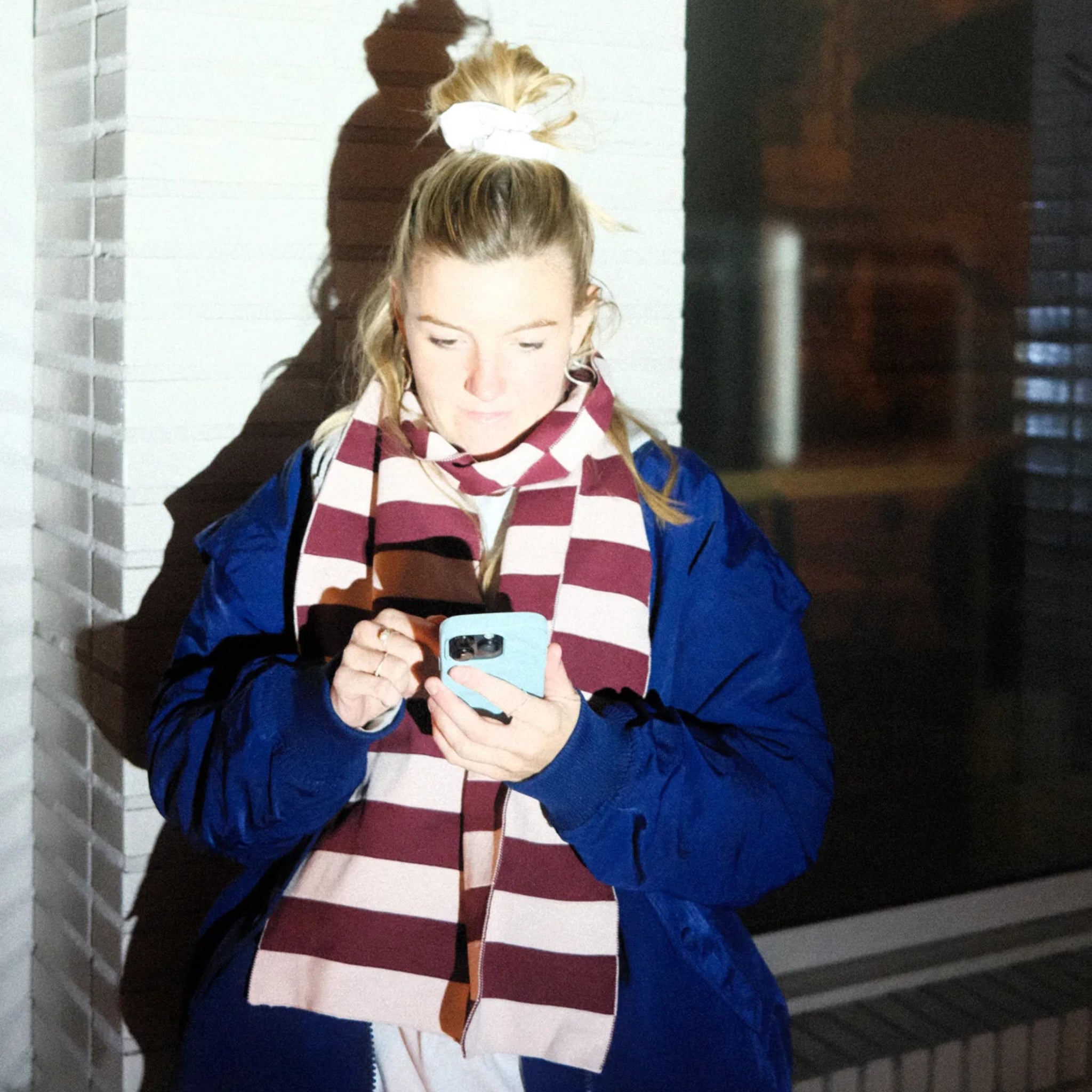 Een persoon met blond haar in een paardenstaart, een blauwe jas aan en de Caro Antwerp Caro Sjaal Ace Pinky Promise, staat tegen een witte bakstenen muur en kijkt naar zijn smartphone terwijl fel licht zijn schaduw werpt.