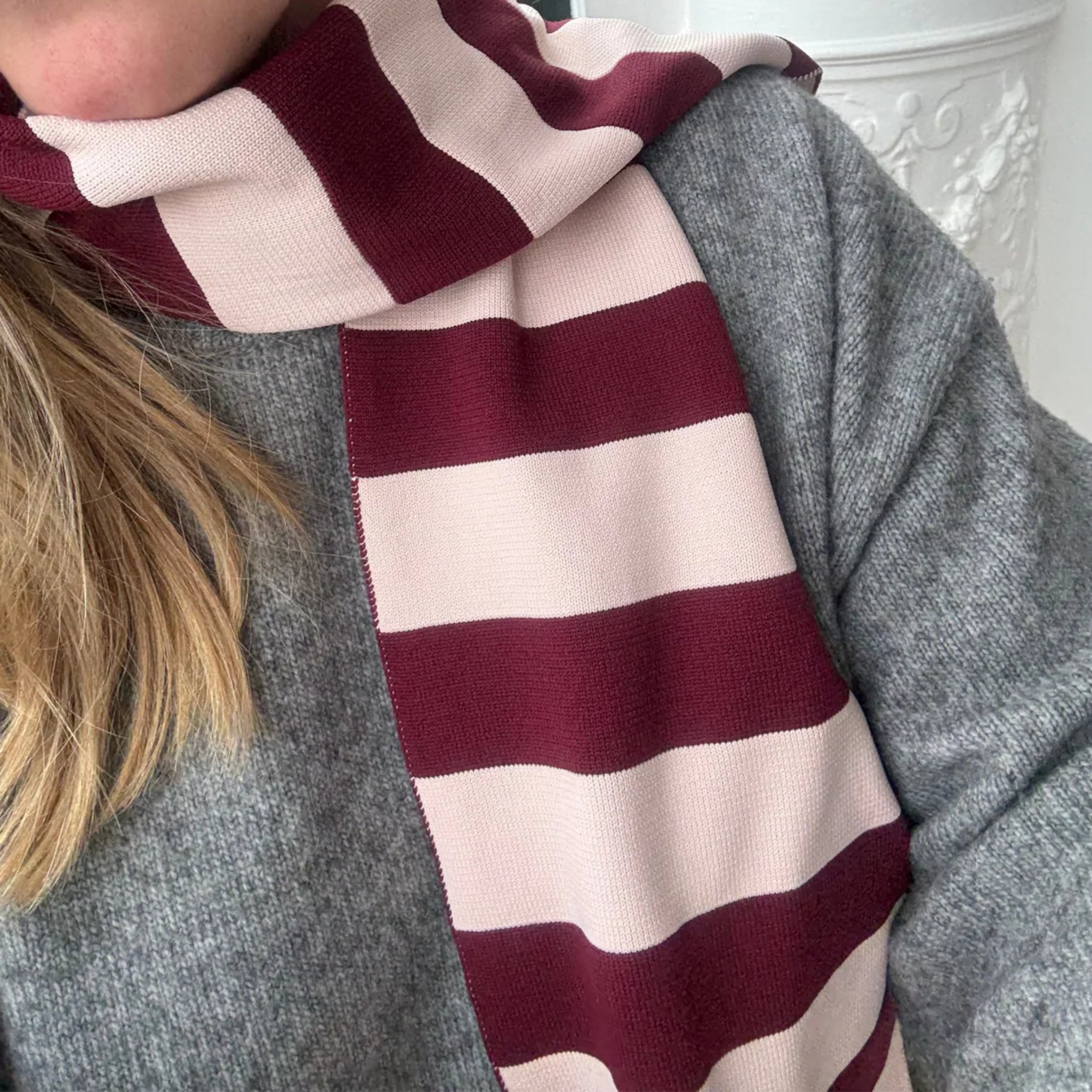 Een persoon draagt een grijze gebreide trui en de Caro Antwerp Sjaal Ace Pinky Promise, met blond haar zichtbaar aan de linkerkant. De witte achtergrond met een decoratief element weerspiegelt de gezellige stijl van Caro Antwerp.