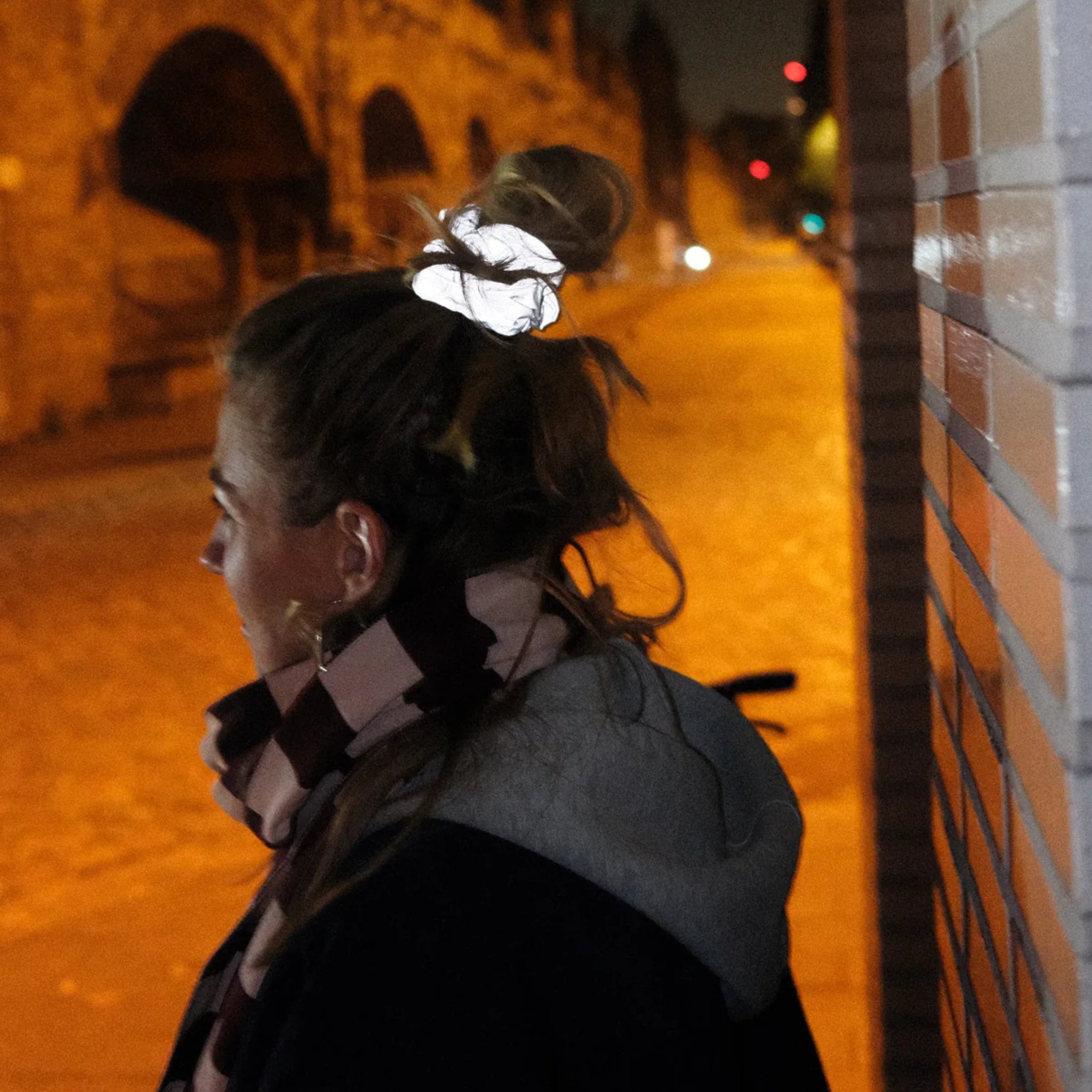 Een persoon met lichtbruin haar in een knot, draagt een Caro Antwerp Caro The Scrunch Club Scrunchie 2 st., staat 's nachts bij een betegelde muur in een schemerige straat, gekleed in een sjaal en jas.
