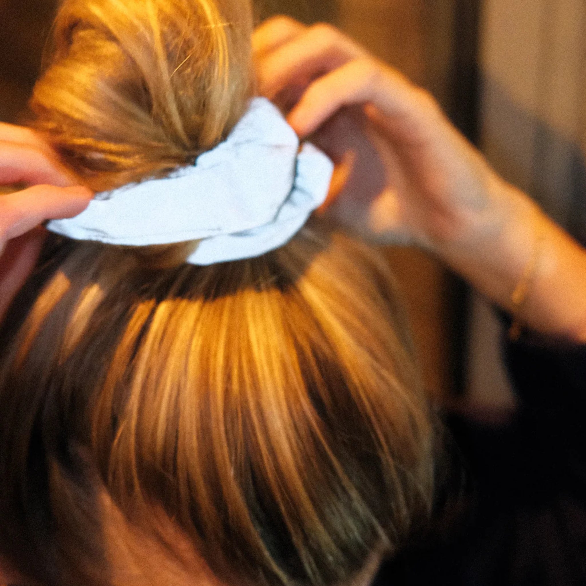 Close-up van iemand die zijn blonde haar met beide handen in een knotje modelleert met behulp van een witte Caro Antwerp Caro The Scrunch Club Scrunchie 2 st.