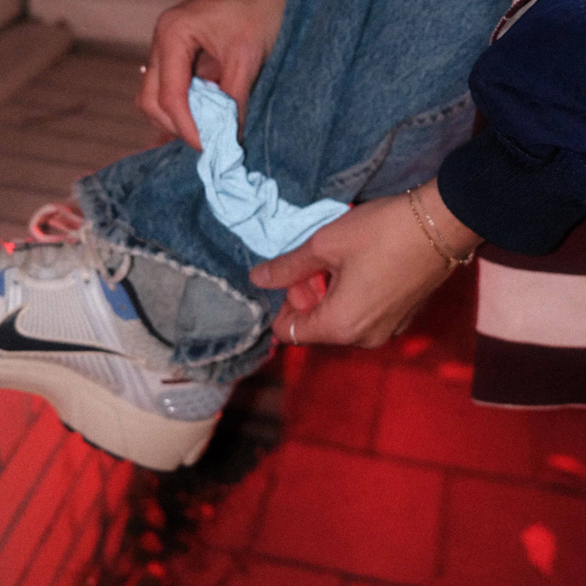 Een persoon in een blauwe spijkerbroek en witte sneakers houdt een Caro Antwerp Caro The Scrunch Club Scrunchie 2 st. bij zijn enkel, oplichtend in rood licht op een stenen stoep - een trendy fietsaccessoire voor elke nachtelijke rit.