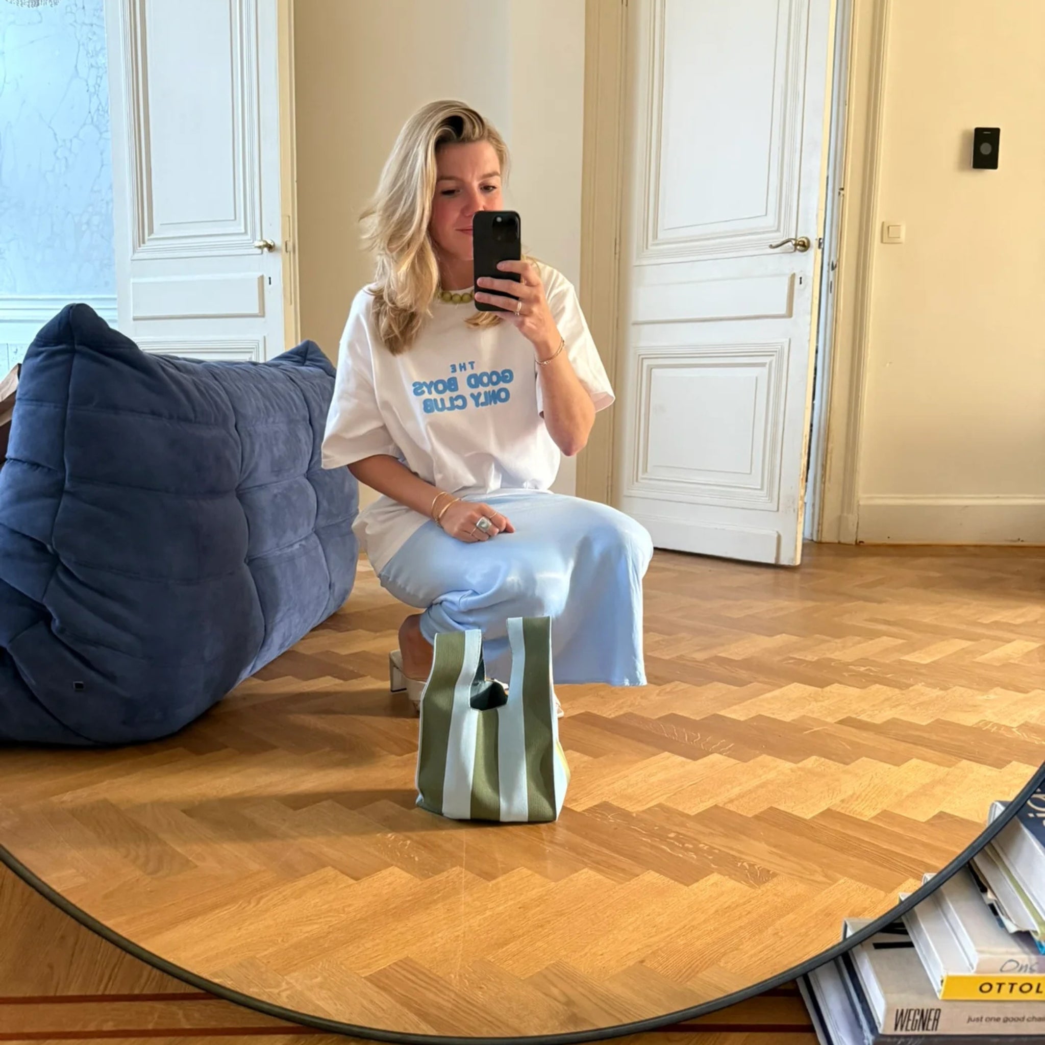 Een vrouw met blond haar in een wit T-shirt en lichtblauwe rok zit op een blauwe loungestoel en neemt een spiegel selfie in een lichte kamer met houten vloeren. Een Caro Antwerp Caro Handtas Stripe Dolce Far Niente Olivia en boeken liggen op de vloer.