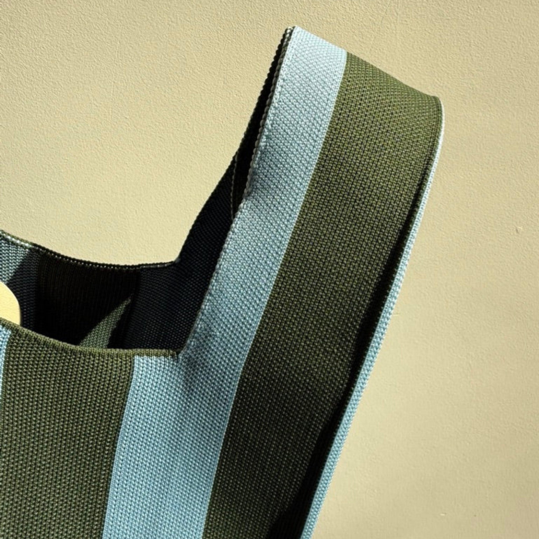 Een close-up van de Caro Handtas Stripe Dolce Far Niente Olivia van Caro Antwerp, met dikke olijfgroene en lichtblauwe strepen tegen een effen beige achtergrond.