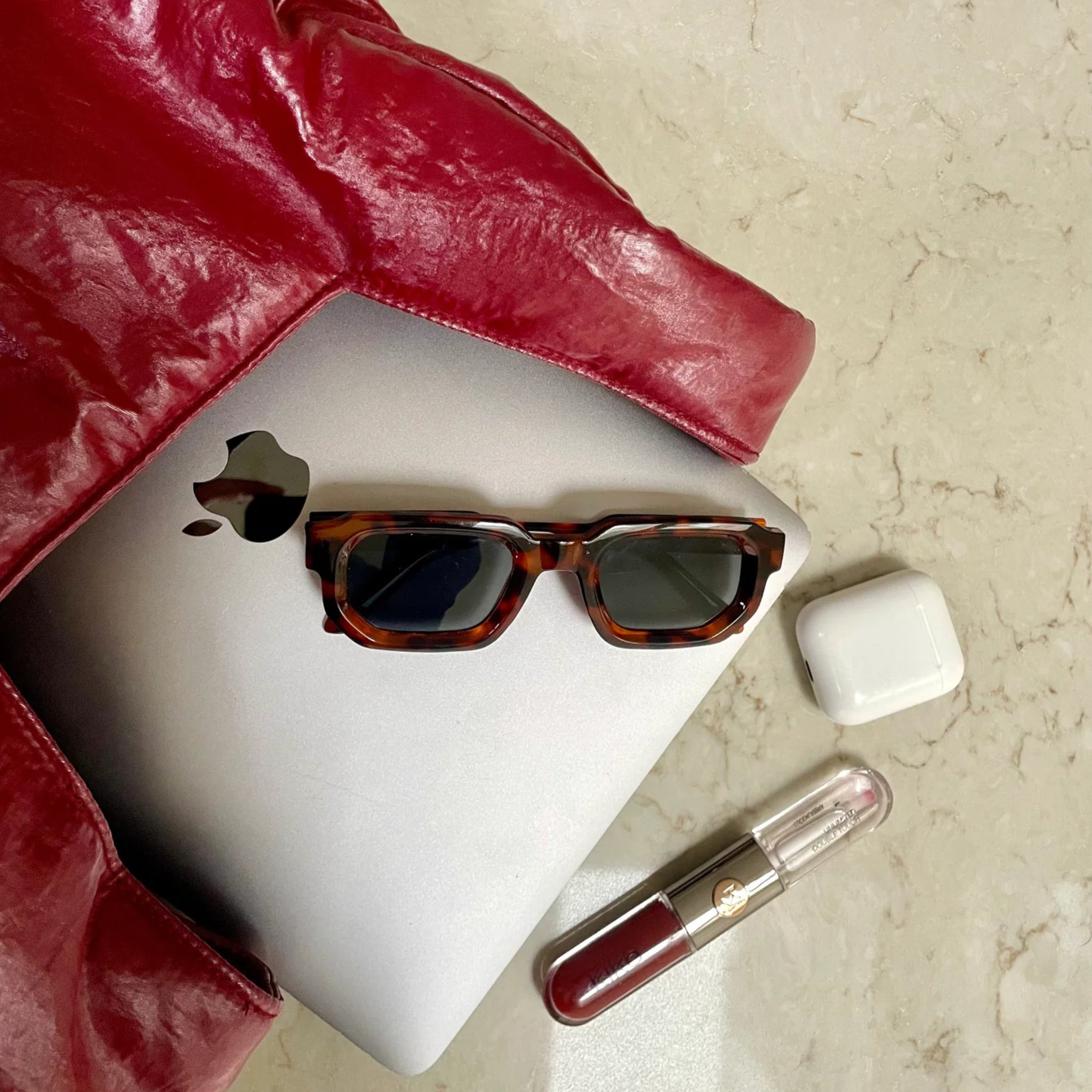 Een zilveren laptop met een zonnebril van schildpad rust gedeeltelijk in een Caro Antwerp Caro Handtas Chubby Barolo Bordeaux pofzak. Op het beige oppervlak liggen witte draadloze oordopjes in een etui en een rode en doorzichtige lipgloss met twee uiteinden.