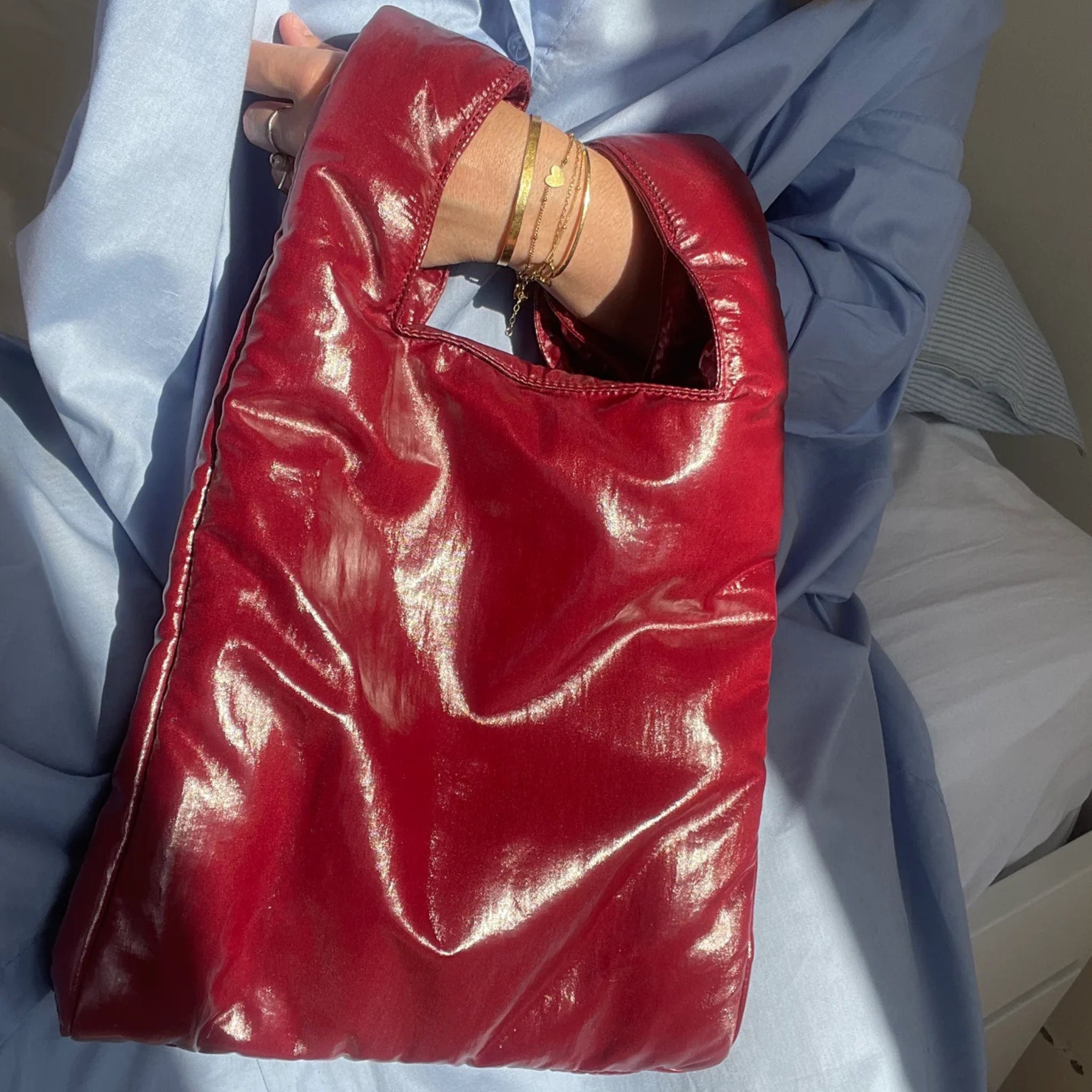 Een persoon in een lichtblauwe outfit houdt de glanzende Caro Antwerp Caro Handtas Chubby Barolo Bordeaux met een vierkant handvat vast en draagt gouden armbanden. Zonlicht benadrukt de glanzende textuur van de pofzak.
