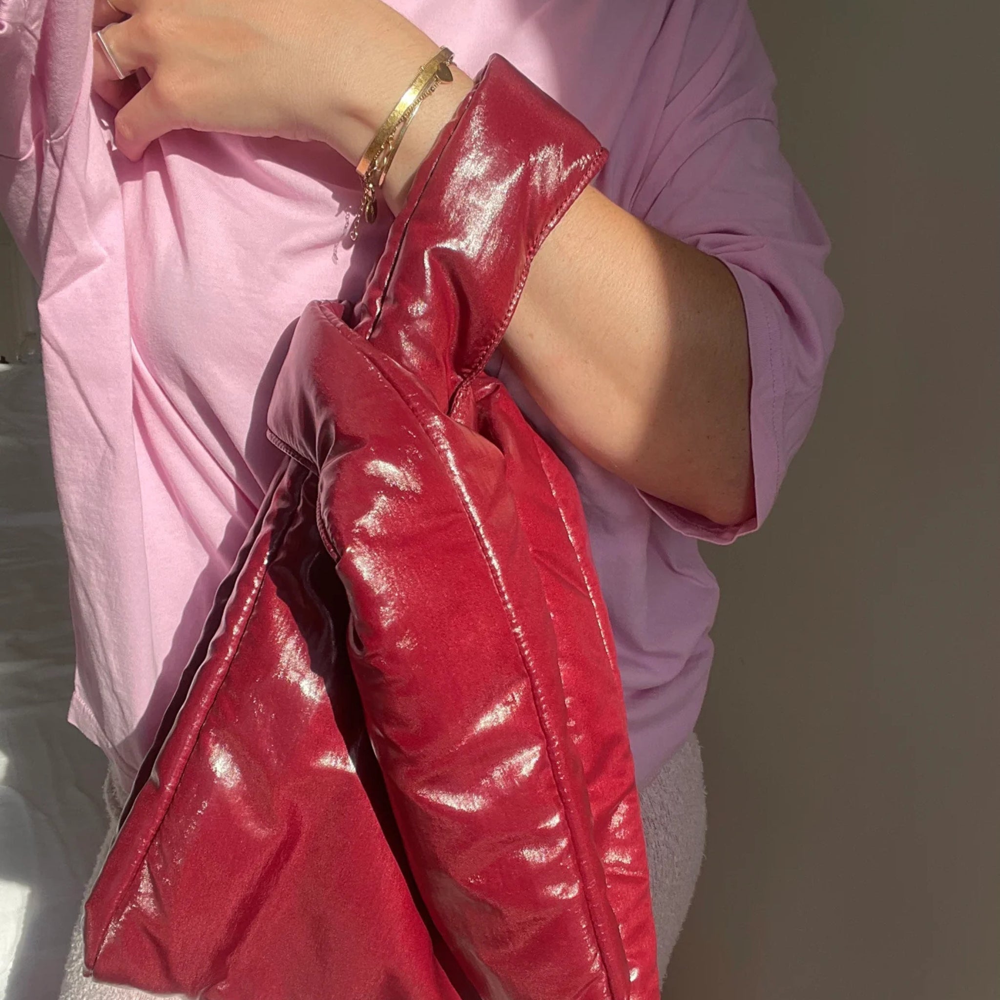 Een persoon in een lichtroze shirt met lange mouwen houdt de Caro Antwerp Caro Handtas Chubby Barolo Bordeaux pofzak met knoopdetail vast. Zonlicht werpt schaduwen op hun arm, waardoor een gouden armband wordt benadrukt tegen een neutrale, zacht verlichte achtergrond.