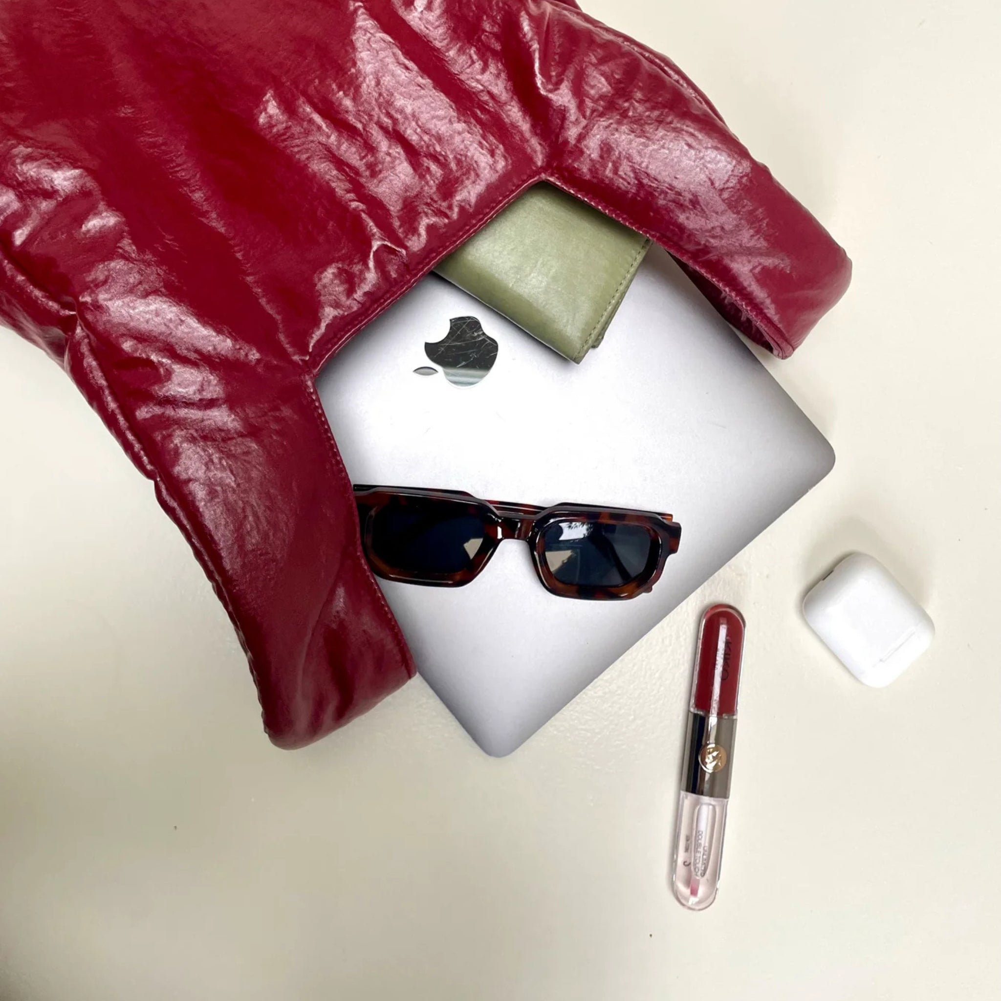 Een Caro Antwerp Handtas Chubby Barolo Bordeaux ligt op een licht oppervlak met een groene portemonnee, een gesloten zilveren MacBook, een donkere zonnebril, rode lippenstift en witte draadloze oordopjes.