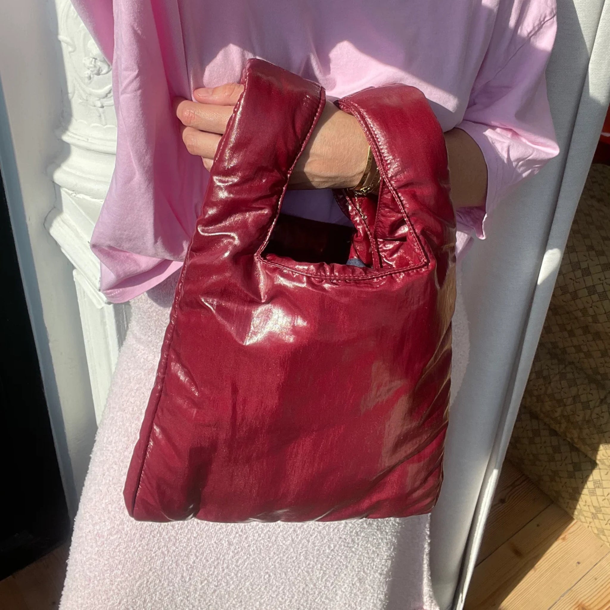 Een persoon in een lichtroze topje houdt de glanzende, gevoerde Caro Handtas Chubby Barolo Bordeaux van Caro Antwerp vast. Zonlicht accentueert de glans van de tas tegen een witte muur en houten vloer achtergrond.
