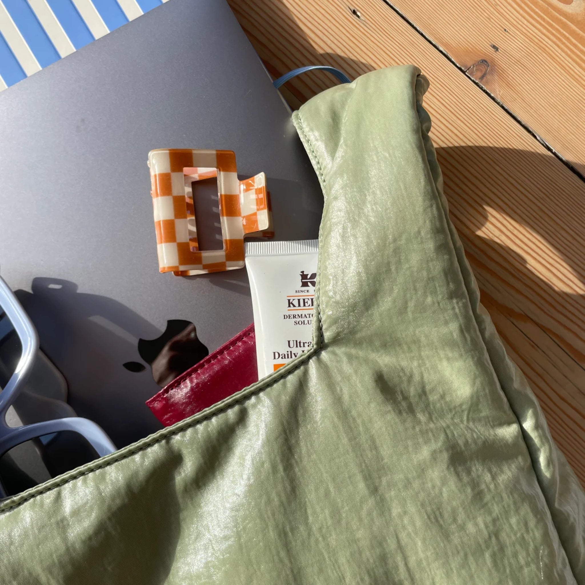 Een Antwerpse Caro Handtas Chubby Matcha Groen rust op een houten oppervlak met een zilveren laptop, een oranje geruite haarspeld, een zonnebril, een rood voorwerp en een tube Kiehl's Ultra Facial Daily UV Defense. Zonlicht werpt zachte schaduwen over de handtas-scène.
