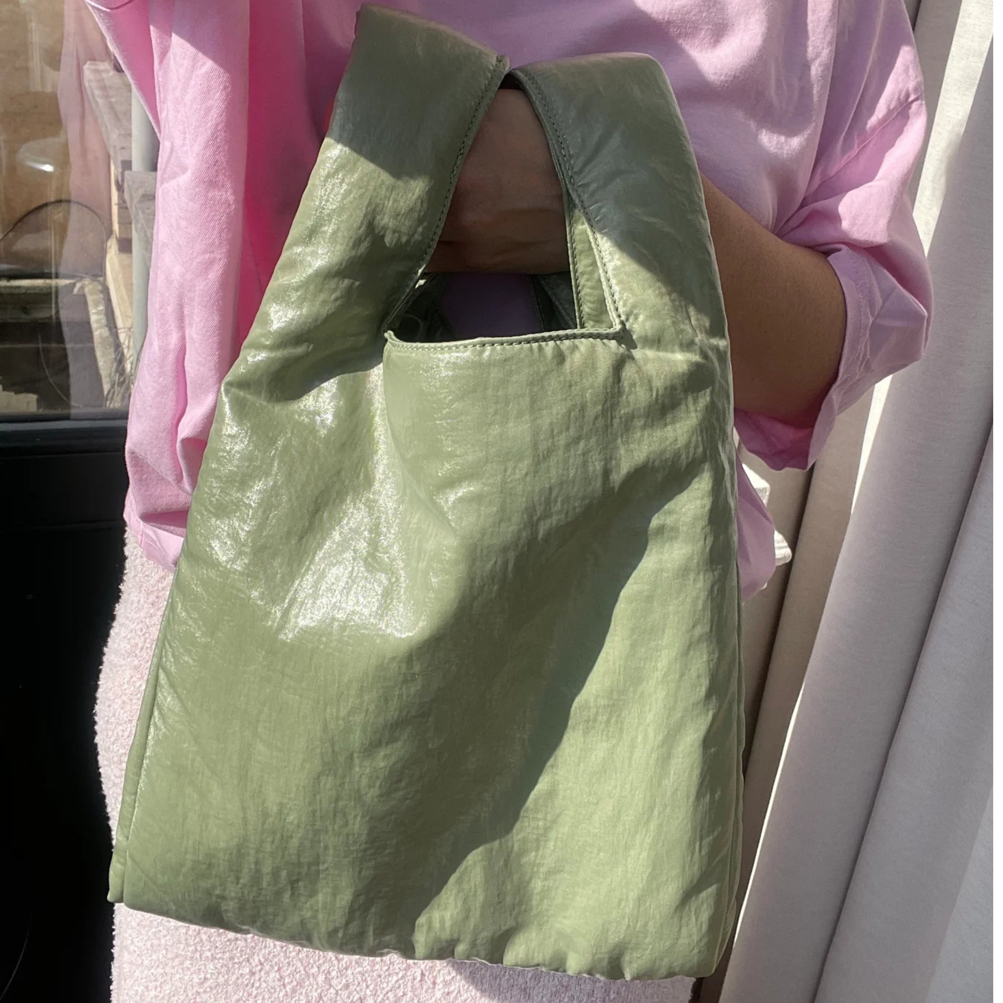 Een persoon met een roze topje houdt een grote, glanzende Caro Antwerp Caro Handtas Chubby Matcha Groen pofzak met een kort hengsel vast en staat bij een raam waar licht naar binnen schijnt.