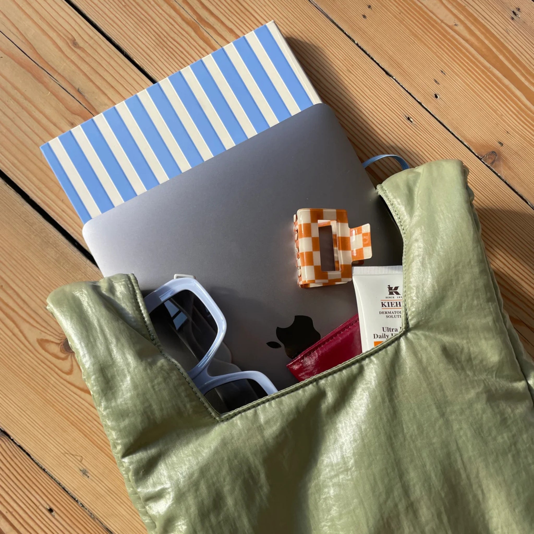 De Caro Antwerp Caro Handtas Chubby Matcha Groen rust op een houten vloer, netjes met een gesloten laptop, een blauw gestreept notitieboekje, een witte zonnebril, een oranje geruite haarspeld, een klein wit lotionflesje en een rood voorwerp, georganiseerd in ware handtassenstijl.