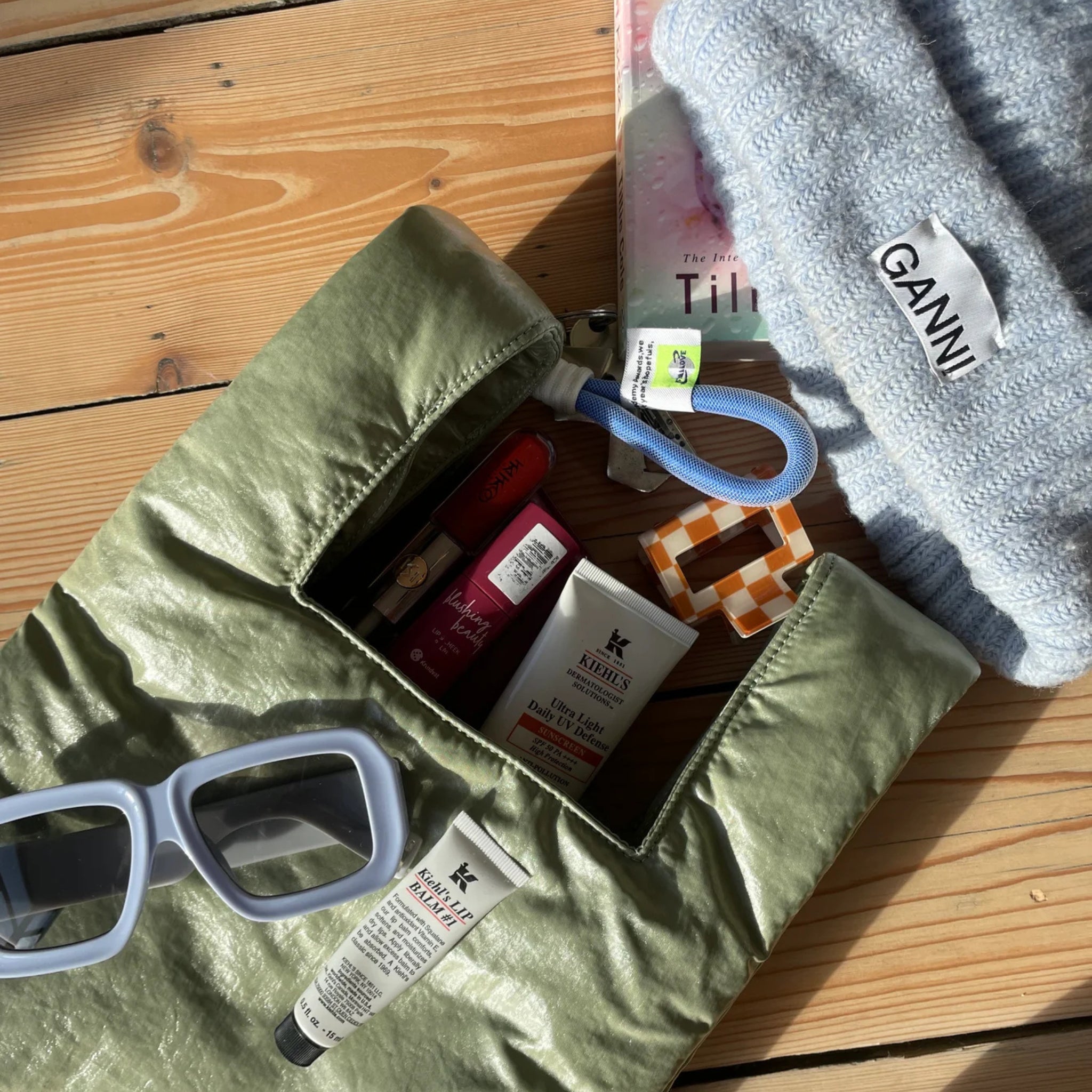 Een Caro Antwerp Handtas Chubby Matcha Groen met zonnebril, lippenbalsem, handcrème, een blauwe GANNI beanie, een boek en een sleutelhanger ligt op een houten oppervlak dat baadt in het zonlicht.