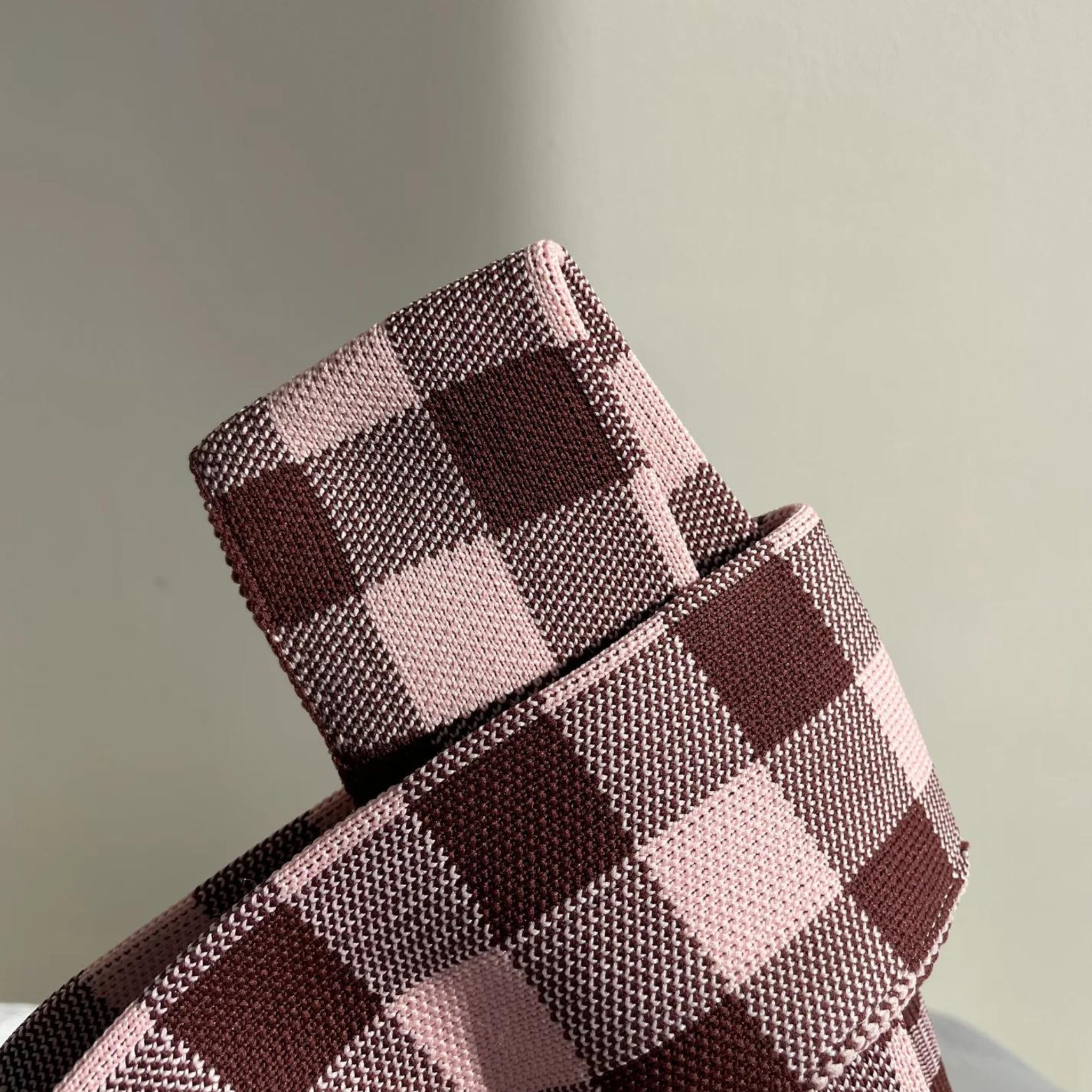 Een close-up van de Caro Antwerp Caro Handtas Les Carreaux Bordeaux Roze, met een ceintuur van structuurstof met een vichyprint in afwisselend lichtroze en donkerbruine vierkantjes tegen een effen lichte achtergrond.