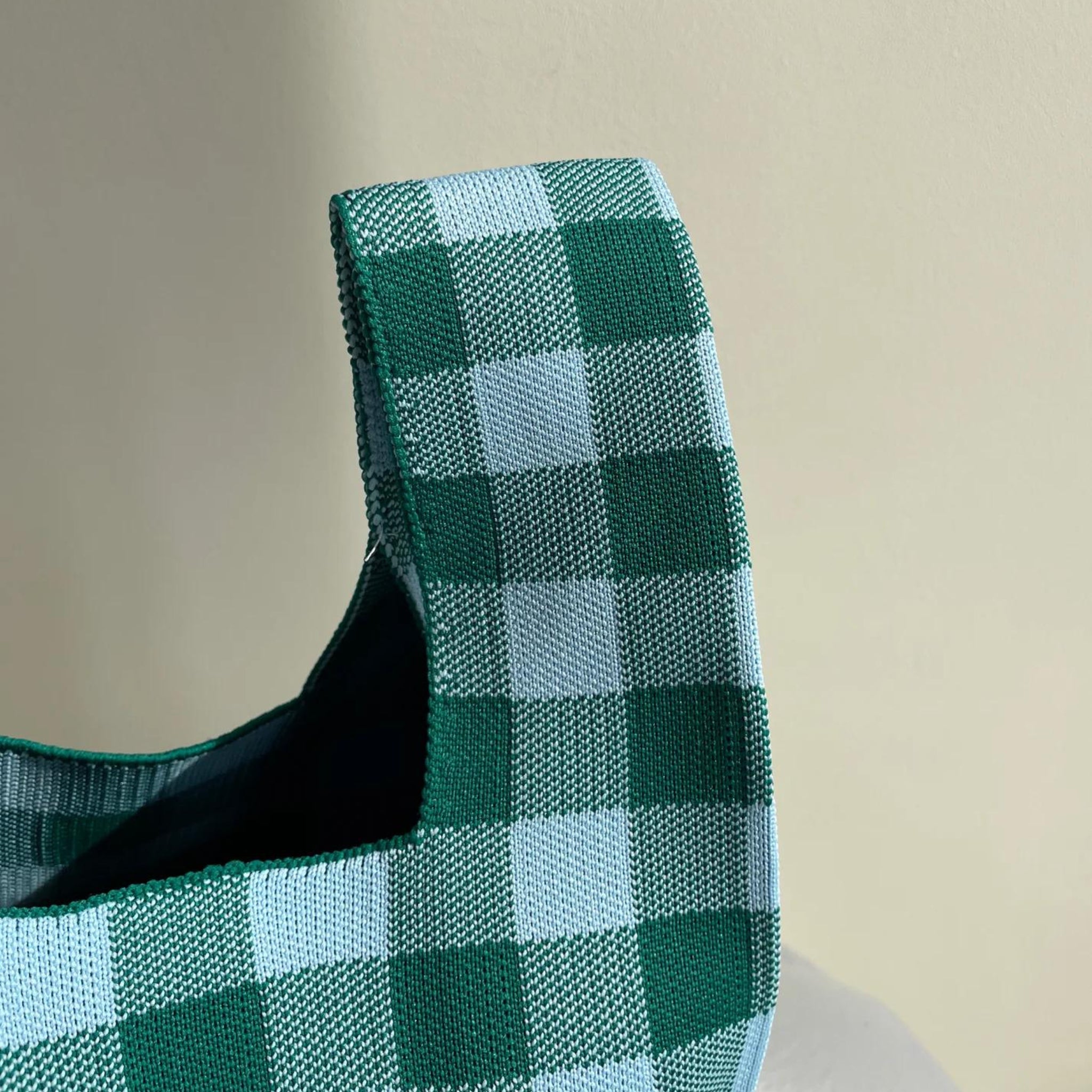 Een close-up van de Caro Handtas Les Carreaux Groen Lichtblauw van Caro Antwerp, met een groen en lichtblauw ruitpatroon, wordt getoond tegen een effen beige achtergrond met zichtbare stoftextuur en stikseldetails.