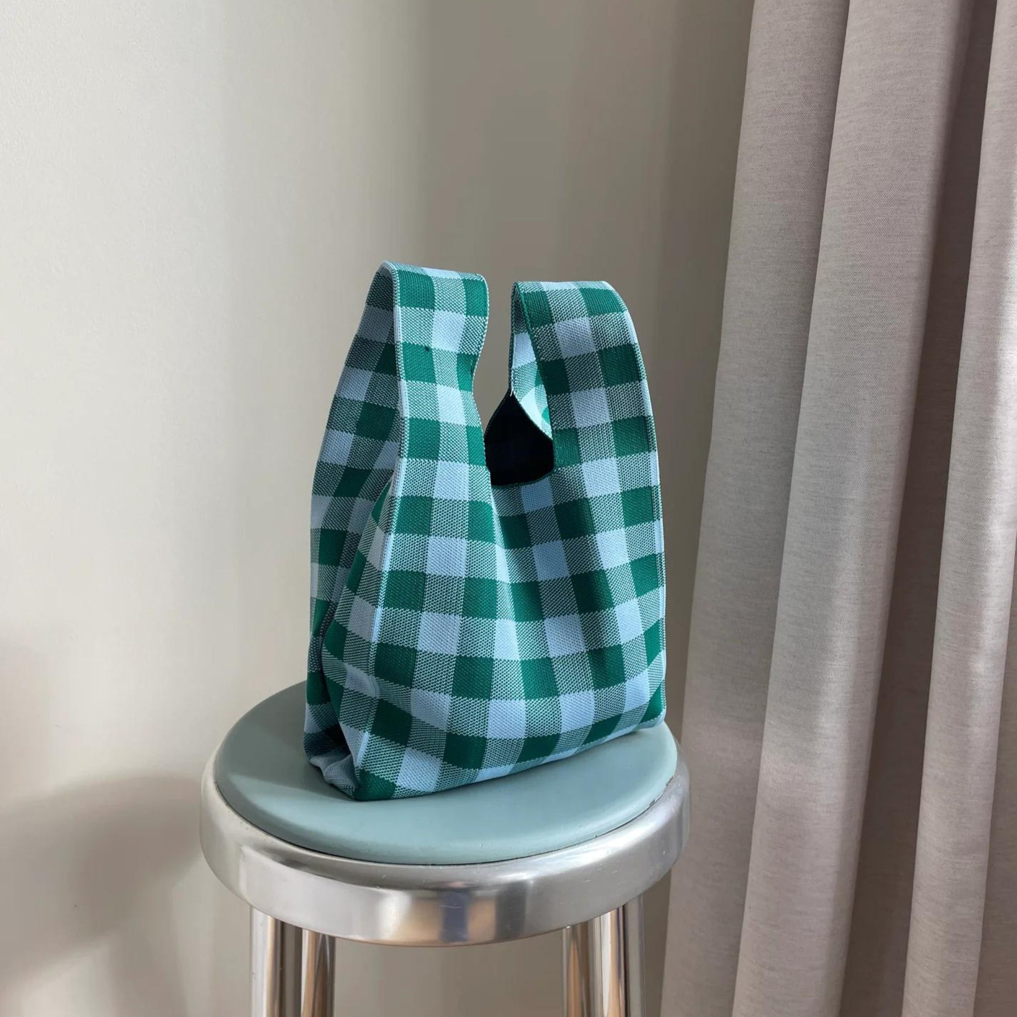 De Caro Antwerp Handtas Les Carreaux Groen Lichtblauw rust op een zilver metalen krukje naast beige gordijnen en een bleke muur.
