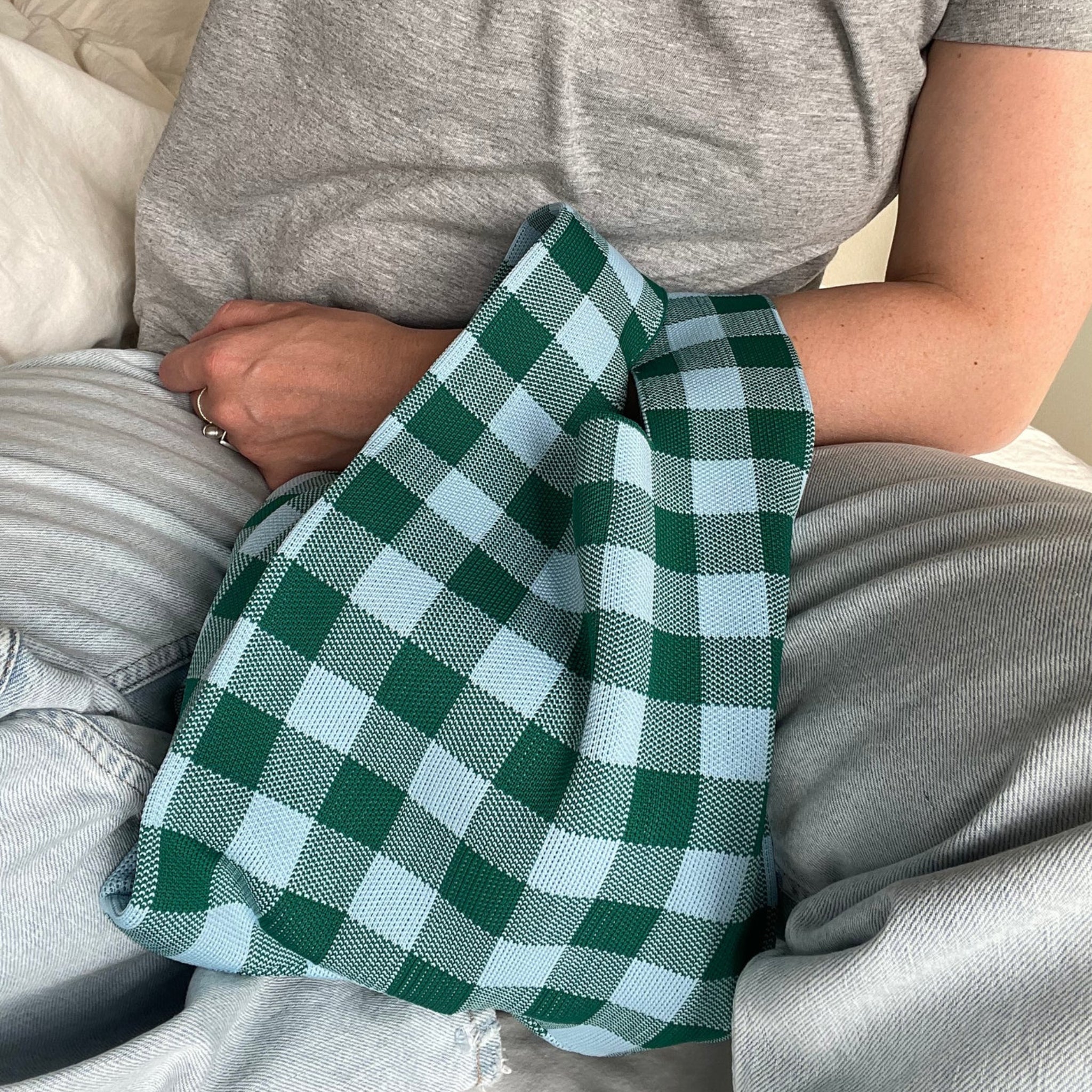 Een persoon in een grijs t-shirt en lichte spijkerbroek heeft een Caro Antwerp Caro Handtas Les Carreaux Groen Lichtblauw op schoot, met een groen en lichtblauw vichy geruit doekje over zijn arm gedrapeerd.