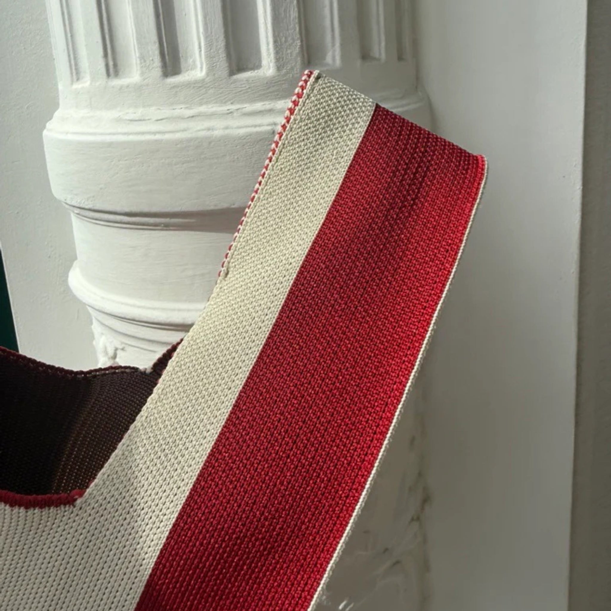 Een close-up van de Caro Handtas Stripe The Casual van Caro Antwerp, met gedurfde rode en beige strepen, gedrapeerd over een witte zuil-een chique geweven accessoire als aanvulling op modieuze kleding.