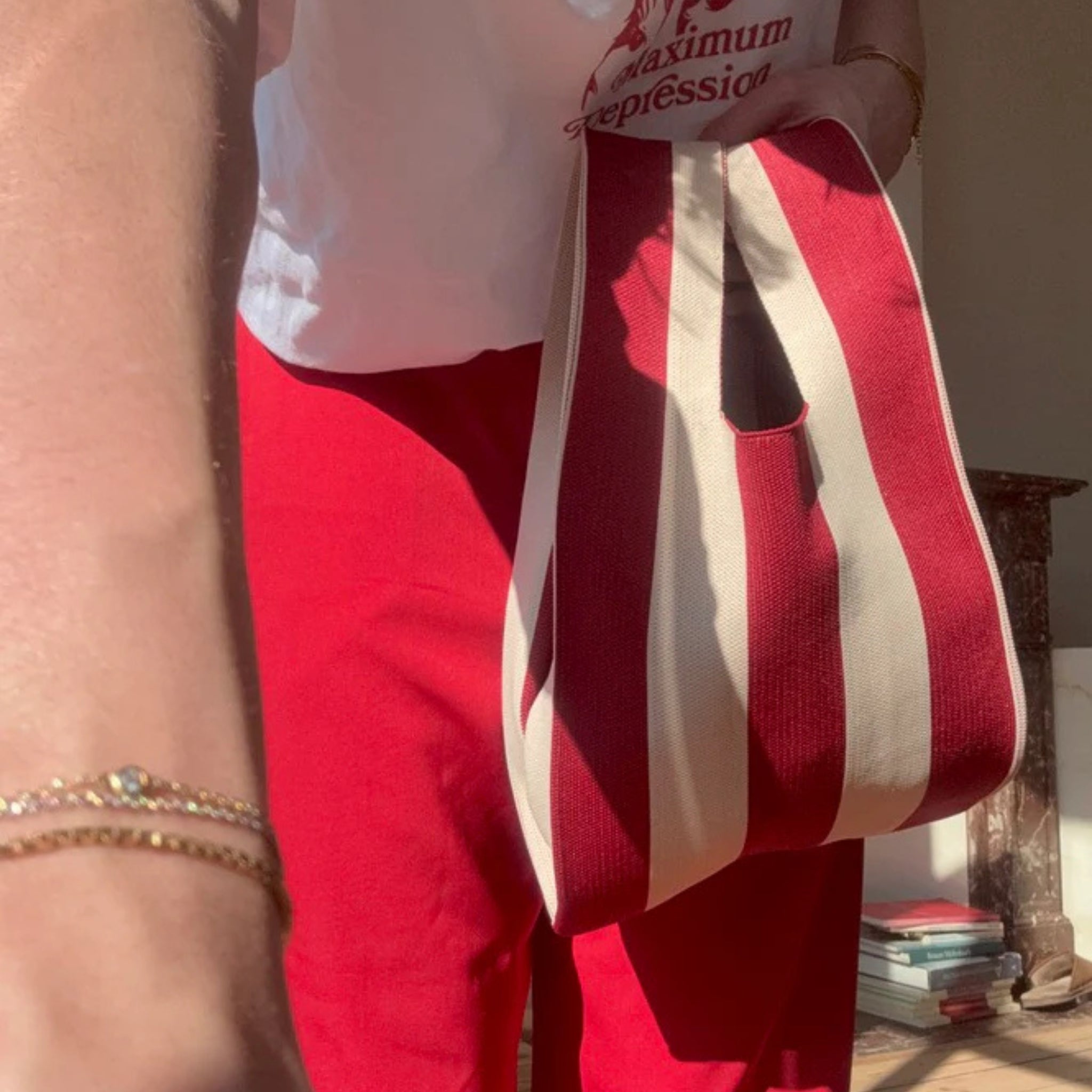 Een persoon in een stijlvolle rode broek en een wit topje houdt de Caro Antwerp Caro Handtas Stripe The Casual vast, met rood-witte strepen. Zonlicht vult de kamer, boeken liggen opgestapeld op de vloer en armbanden glinsteren aan hun arm.