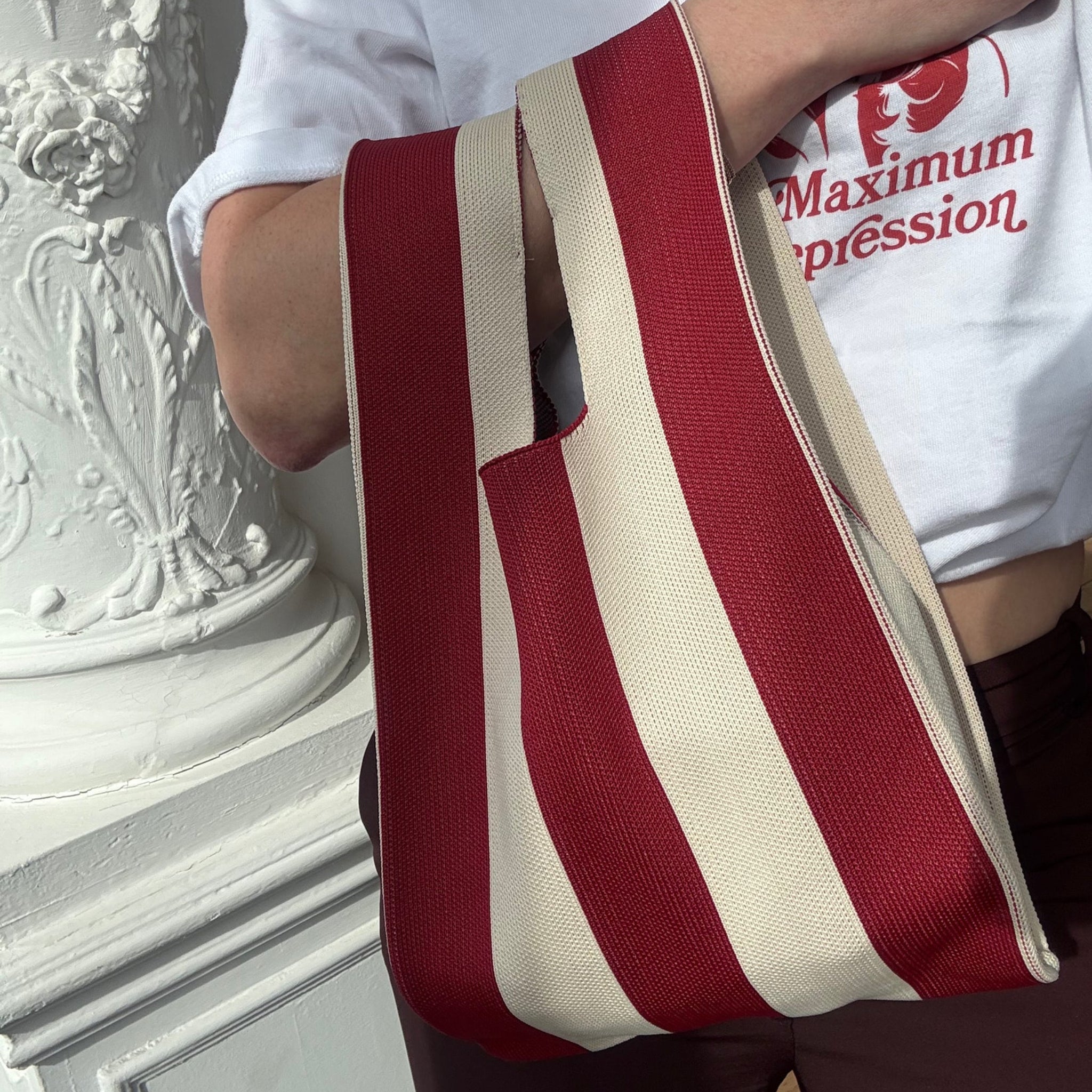 Een persoon houdt de Caro Antwerp Caro Handtas Stripe The Casual, met rode en beige strepen, vast terwijl hij een wit shirt met rode tekst draagt naast een sierlijke witte zuil.