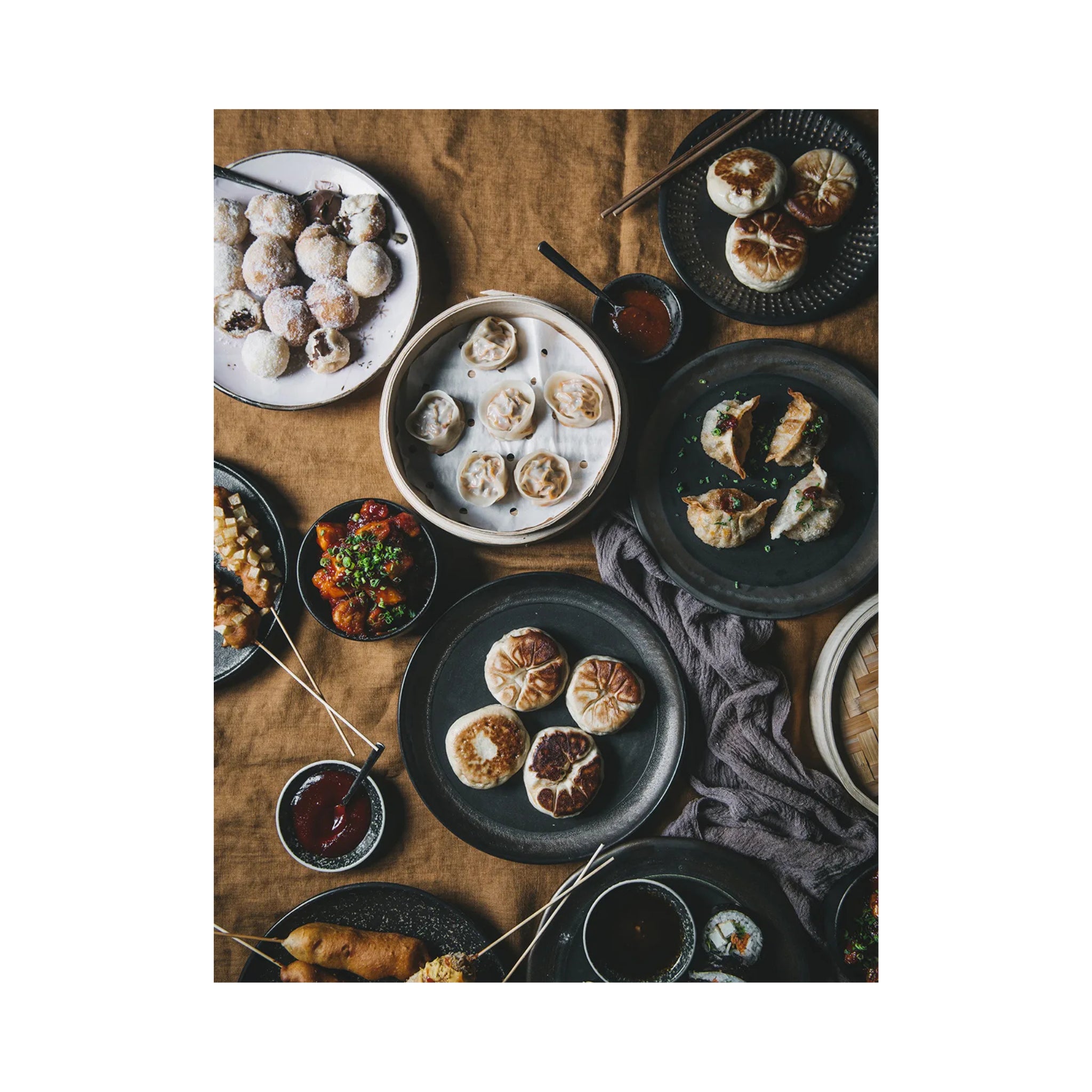 Een bovenaanzicht van geassorteerde dim sum en bao buns - de perfecte inspiratie uit het Cozy Publishing Menu Recepten Voor Gedeelde Momenten Kookboek - mooi gerangschikt op zwarte borden met dipsauzen op een bruin tafelkleed en linnen servet.