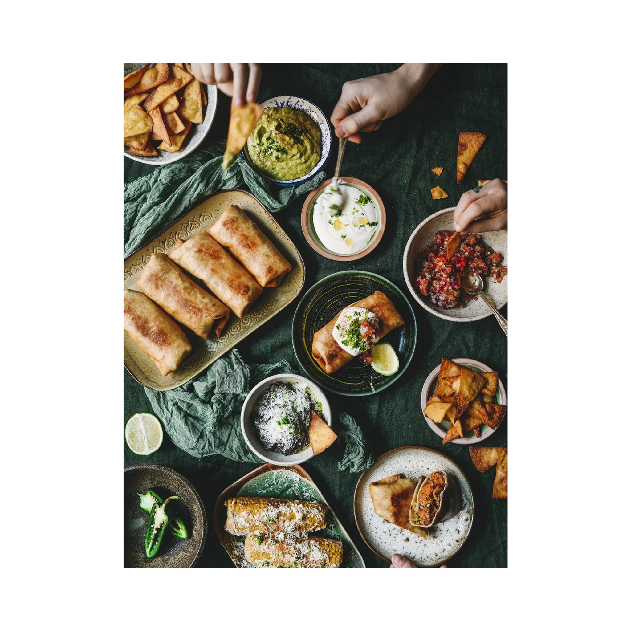 Een bovenaanzicht van een tafel met burrito's, maïs, salsa, guacamole, chips en dipsausjes. Handen reiken naar binnen om het eten te serveren - de levendige essentie van Cozy Publishing Menu Recepten Voor Gedeelde Momenten Kookboek.