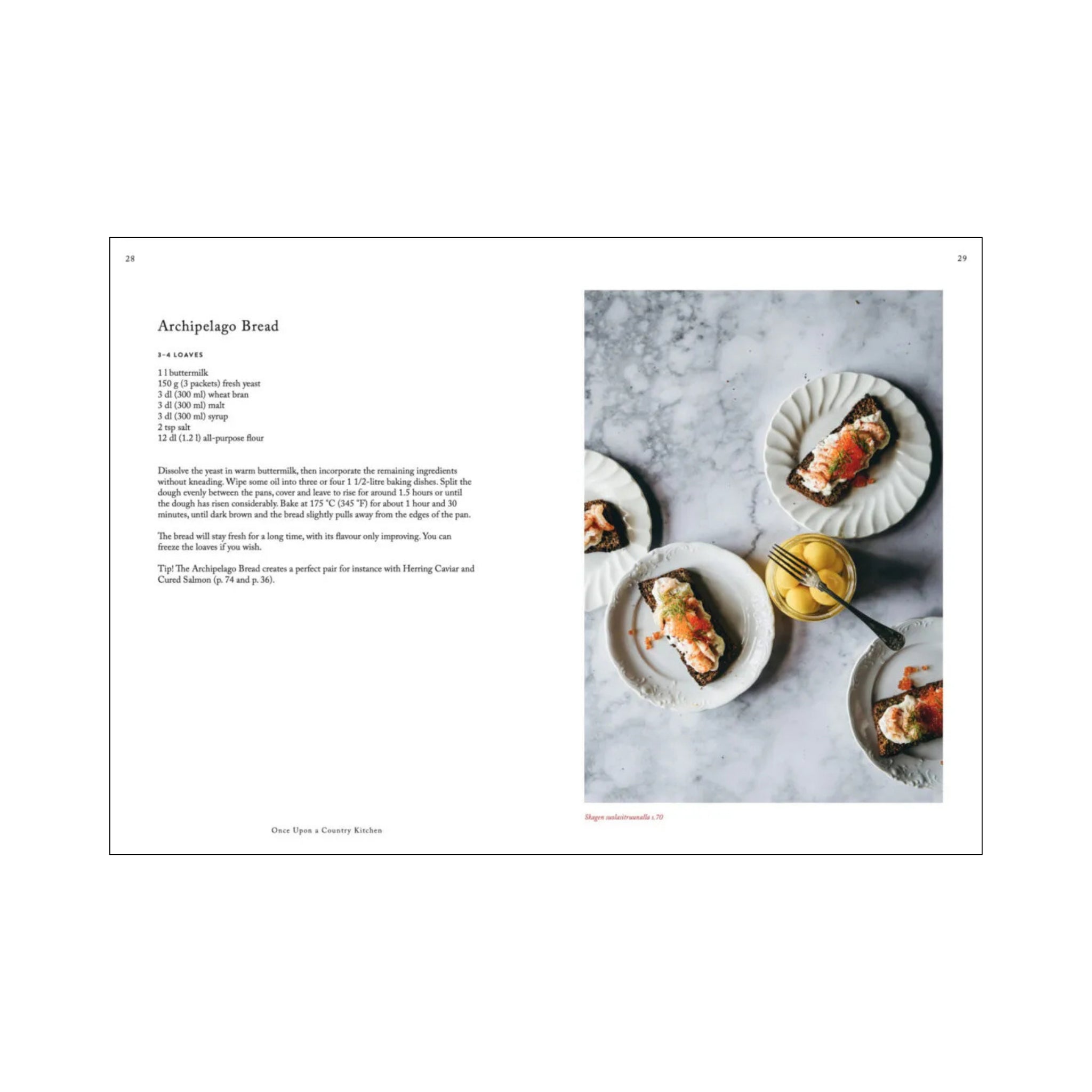 Het Cozy Publishing Once Upon A Country Kitchen Kookboek ligt open voor een broodrecept, terwijl op een marmeren oppervlak boerenborden staan - brood belegd met gerookte zalm en garnering - naast een kleine gouden theepot en kopje.