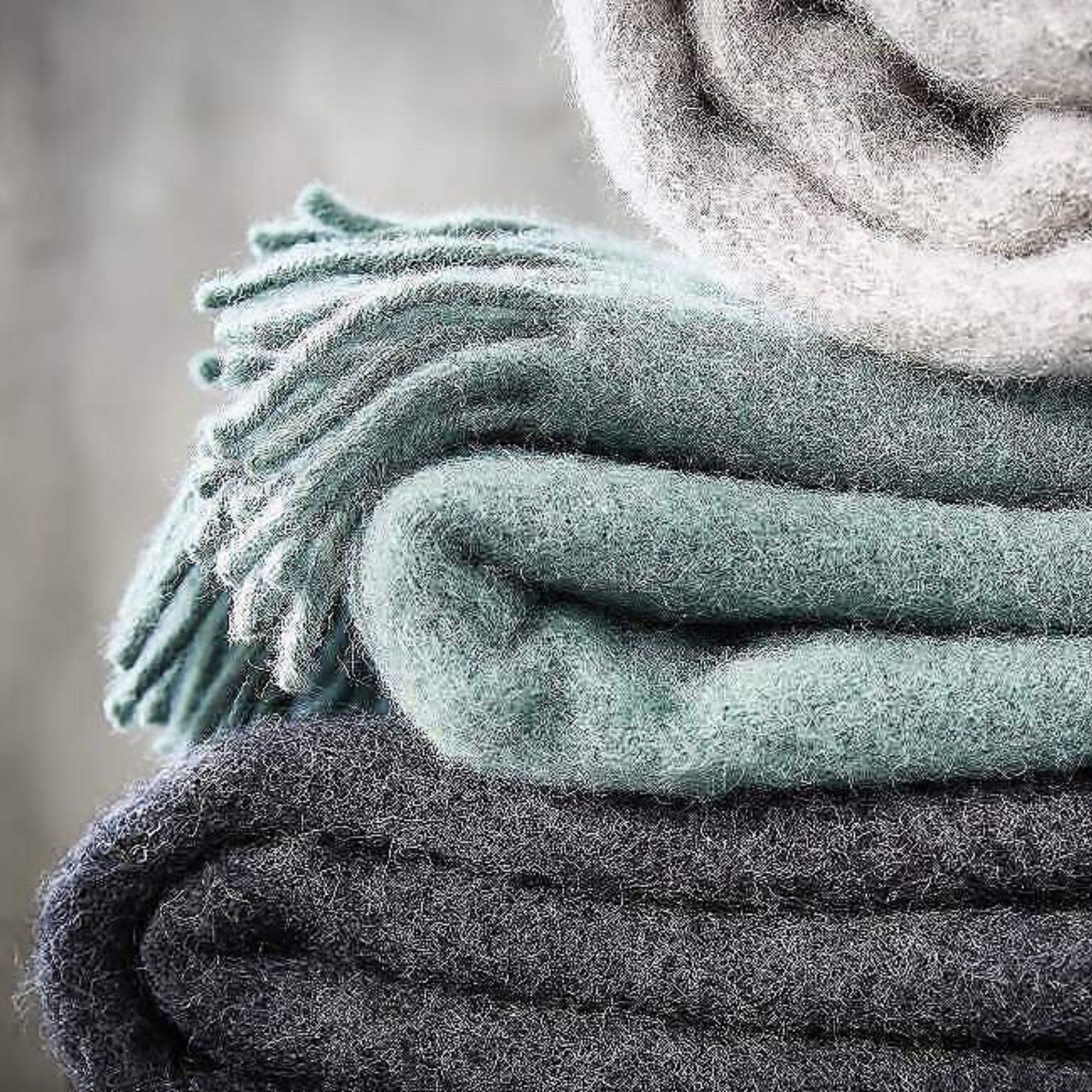 Een close-up van drie netjes gevouwen D&T Design Plaid Wol Antraciet 130/180 dekens, gestapeld in grijs, blauw en lichtgrijs. De bovenste deken heeft franjes aan één rand en de D&T Design textuur ziet er onweerstaanbaar zacht en donzig uit.