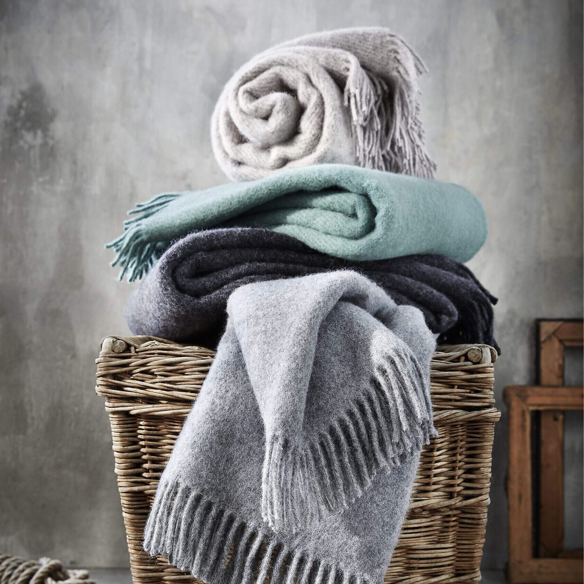 In een rieten mand liggen vier D&T Design Plaid Wol Antraciet 130/180 premium scheerwol dekens in lichtgrijs, donkergrijs, mintgroen en beige, allemaal netjes opgerold en gevouwen tegen een effen grijze muur.