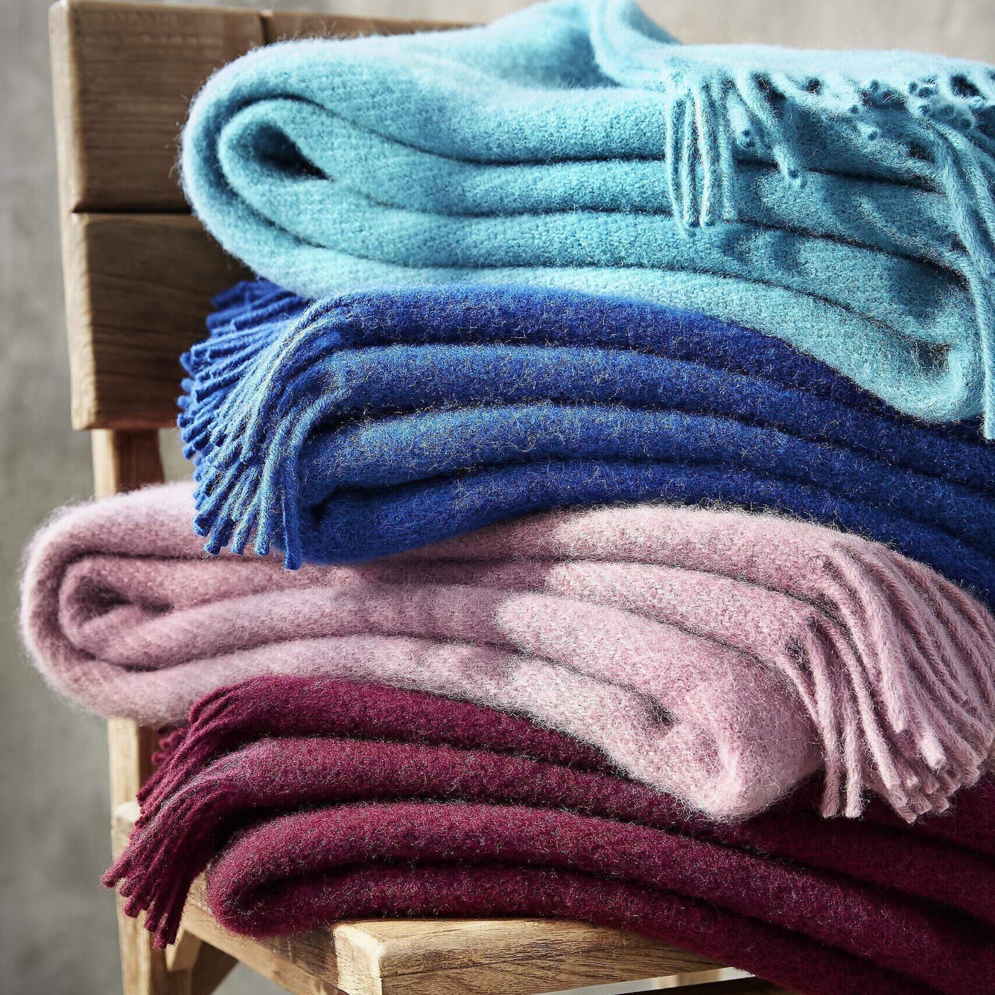 Een stapel van vier D&T Design Plaid Wol Blauw 130/180 dekens in lichtblauw, blauw, roze en bordeaux zuiver wol rust op een houten stoel, met zichtbare franjes aan de randen.