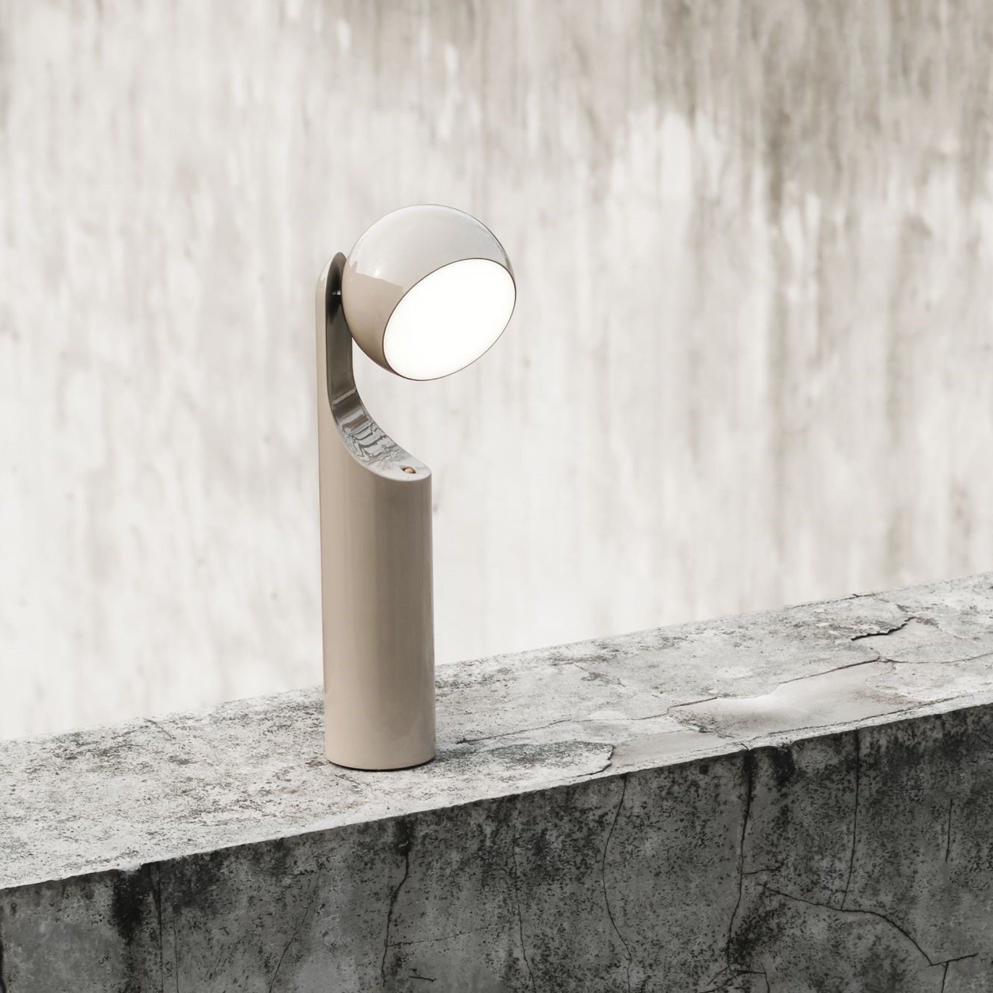 De Fine Lumens Mono Leeslamp LED Oplaadbaar Beige van Fine Lumens, een minimalistische lamp met een afgeronde lamp en beige voet, staat op een verweerde betonnen richel tegen een grijze muur met textuur.