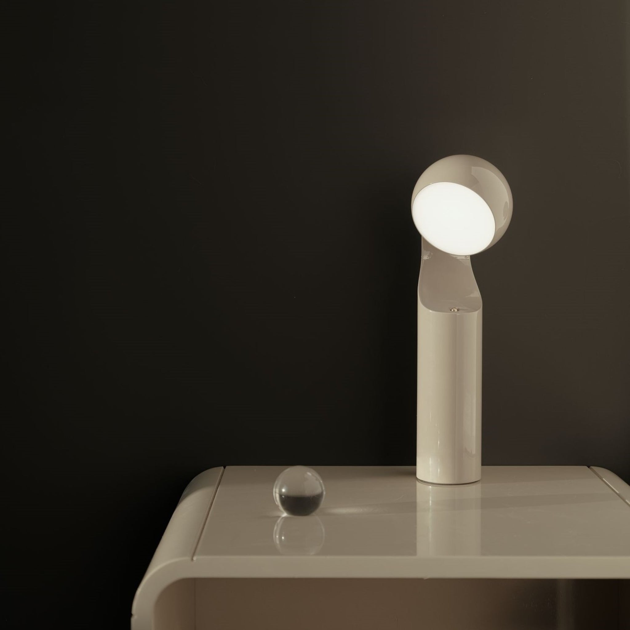 De Fine Lumens Mono Leeslamp LED Oplaadbaar Beige van Fine Lumens staat op een beige nachtkastje tegen een donkere muur, het minimalistische ontwerp en de dimbare LED werpen zacht licht. Vlakbij maakt een kleine glazen bol het vredige tafereel compleet.