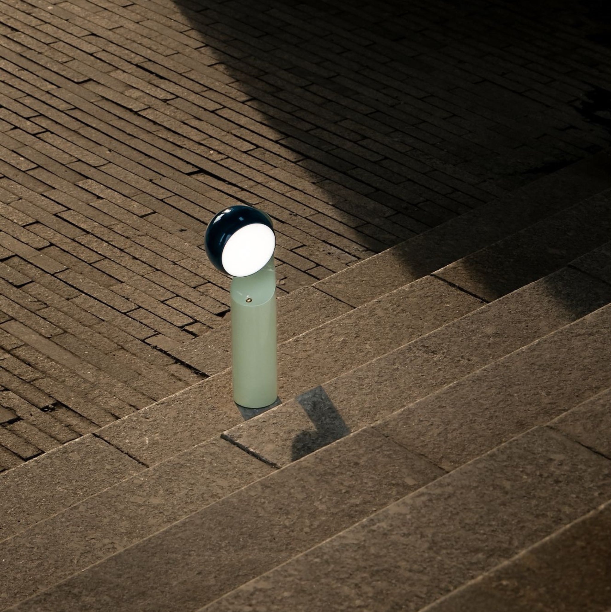 De Fine Lumens Mono Leeslamp LED Oplaadbaar Blauw Munt van Fine Lumens staat naast een stenen trap, zijn gloeiende witte dimbare LED-lamp werpt schaduwen over de gedessineerde bakstenen en stenen buitenoppervlakken.