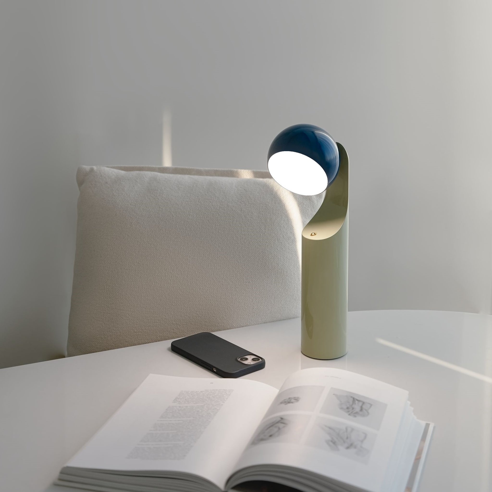 De Fine Lumens Mono Leeslamp LED Oplaadbaar Blauw Munt, met zijn blauwe en witte bolvormige bovenkant, staat op een witte tafel bij een open boek en smartphone, een mix van moderne vorm en functie van Fine Lumens.