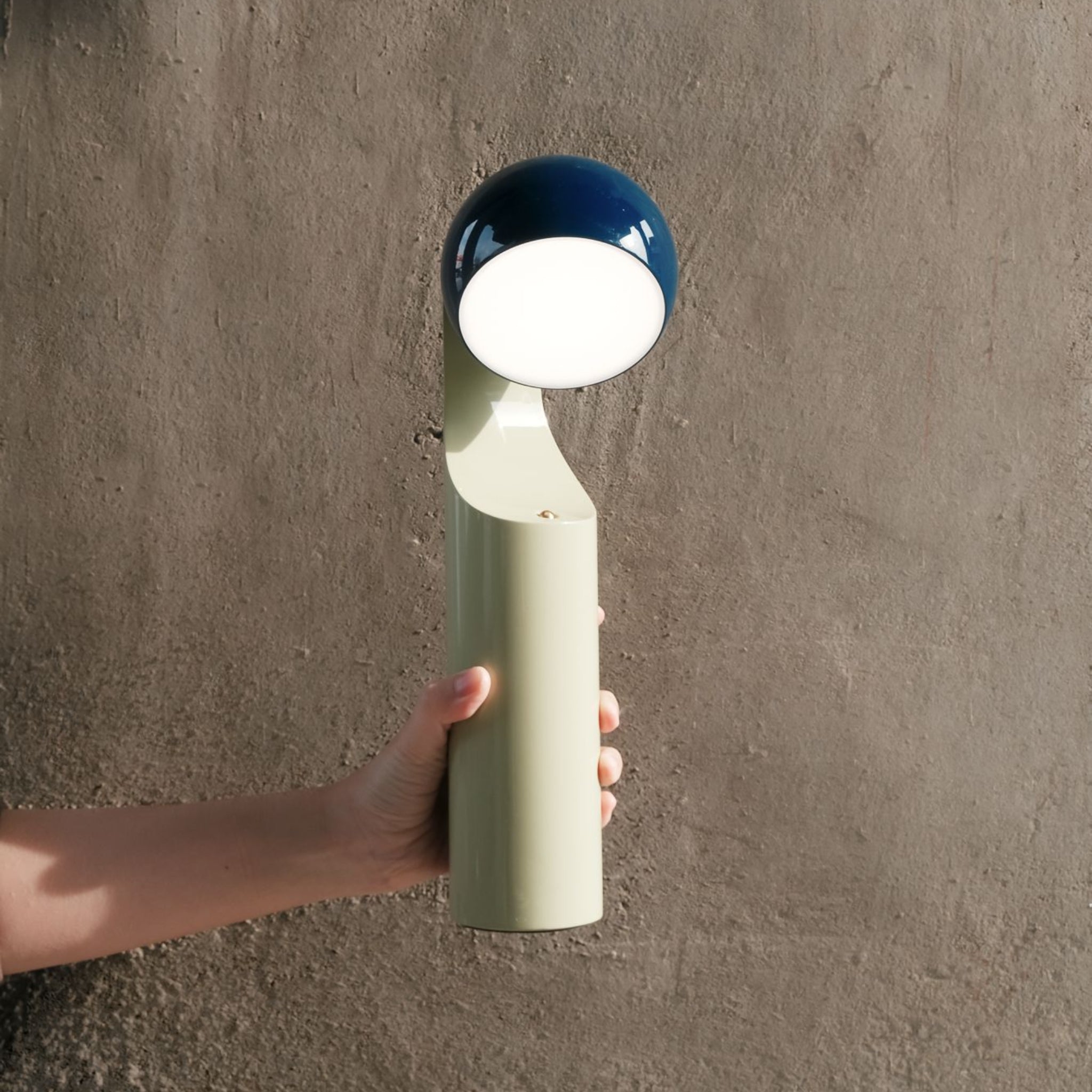 Een hand houdt de Fine Lumens Mono Leeslamp LED Oplaadbaar Blauw Munt vast, een moderne, minimalistische draagbare lamp met een lichtgroene voet en blauwe kop van Fine Lumens, die dimbaar LED-licht uitstraalt tegen een grijze muur met textuur.