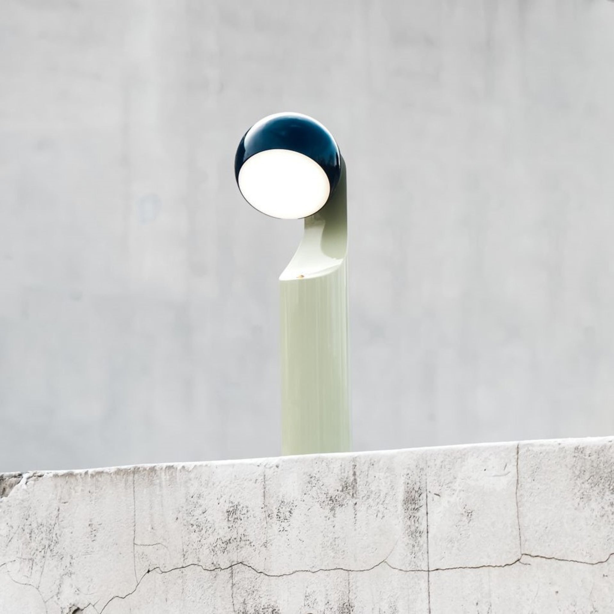 De Fine Lumens Mono Leeslamp LED Oplaadbaar Blauw Munt van Fine Lumens, met een gebogen lichtgroene standaard en dimbare LED-kop, rust op gebarsten beton tegen een effen grijze muur voor een minimalistische look.