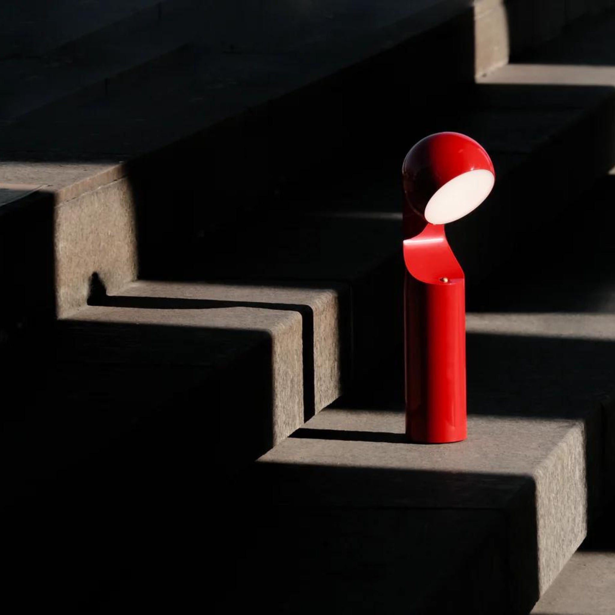 De Fine Lumens Mono Leeslamp LED Oplaadbaar Bright Rood, een dimbare moderne lamp met een afgeronde kop, van Fine Lumens is geplaatst op betonnen traptreden en verlicht een donkere, schaduwrijke ruimte.