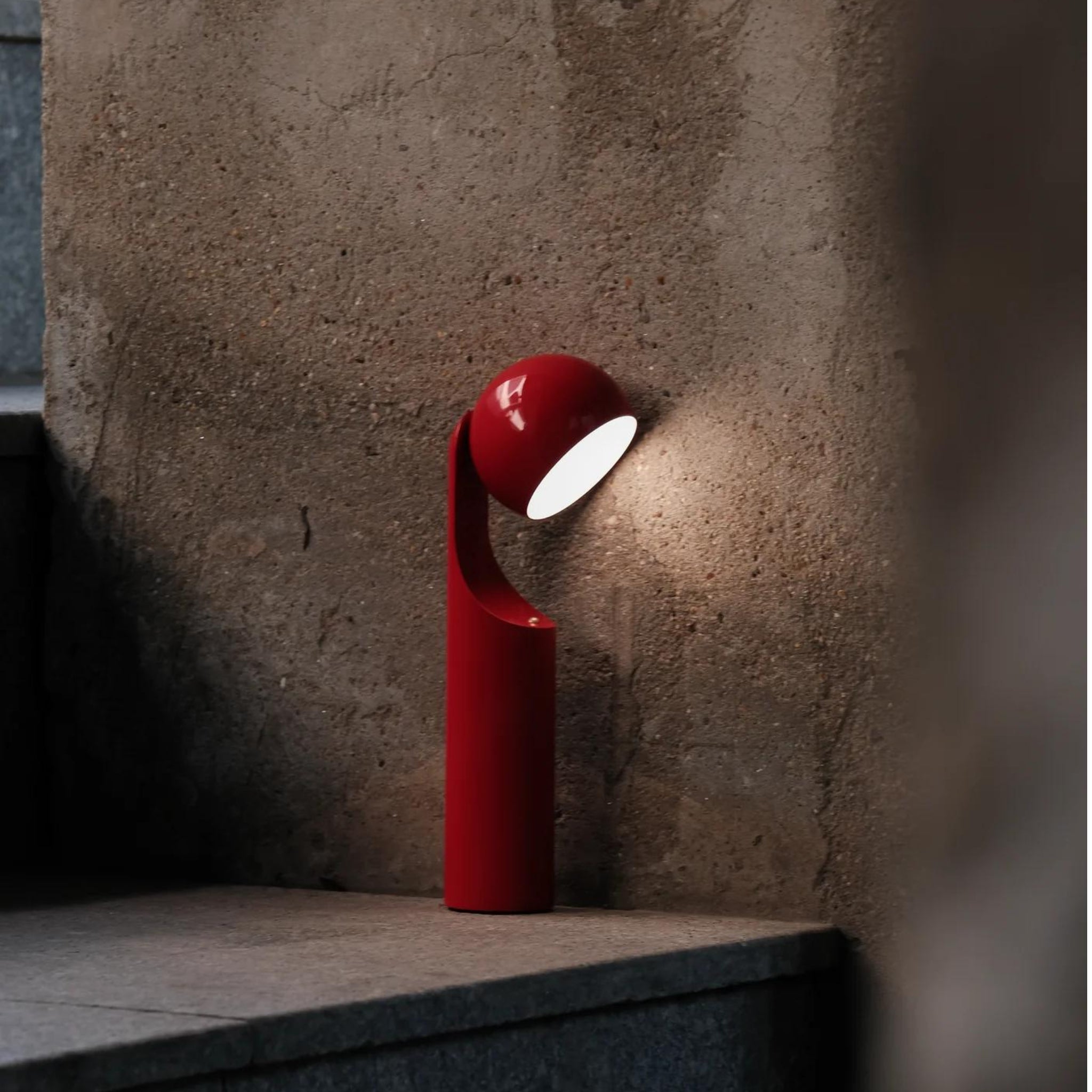 De Fine Lumens Mono Leeslamp LED Oplaadbaar Bright Rood, met een minimalistisch gebogen ontwerp, staat op een stenen vloer bij een beige muur met textuur van Notion Studio en verlicht het oppervlak zachtjes in een schemerige omgeving.