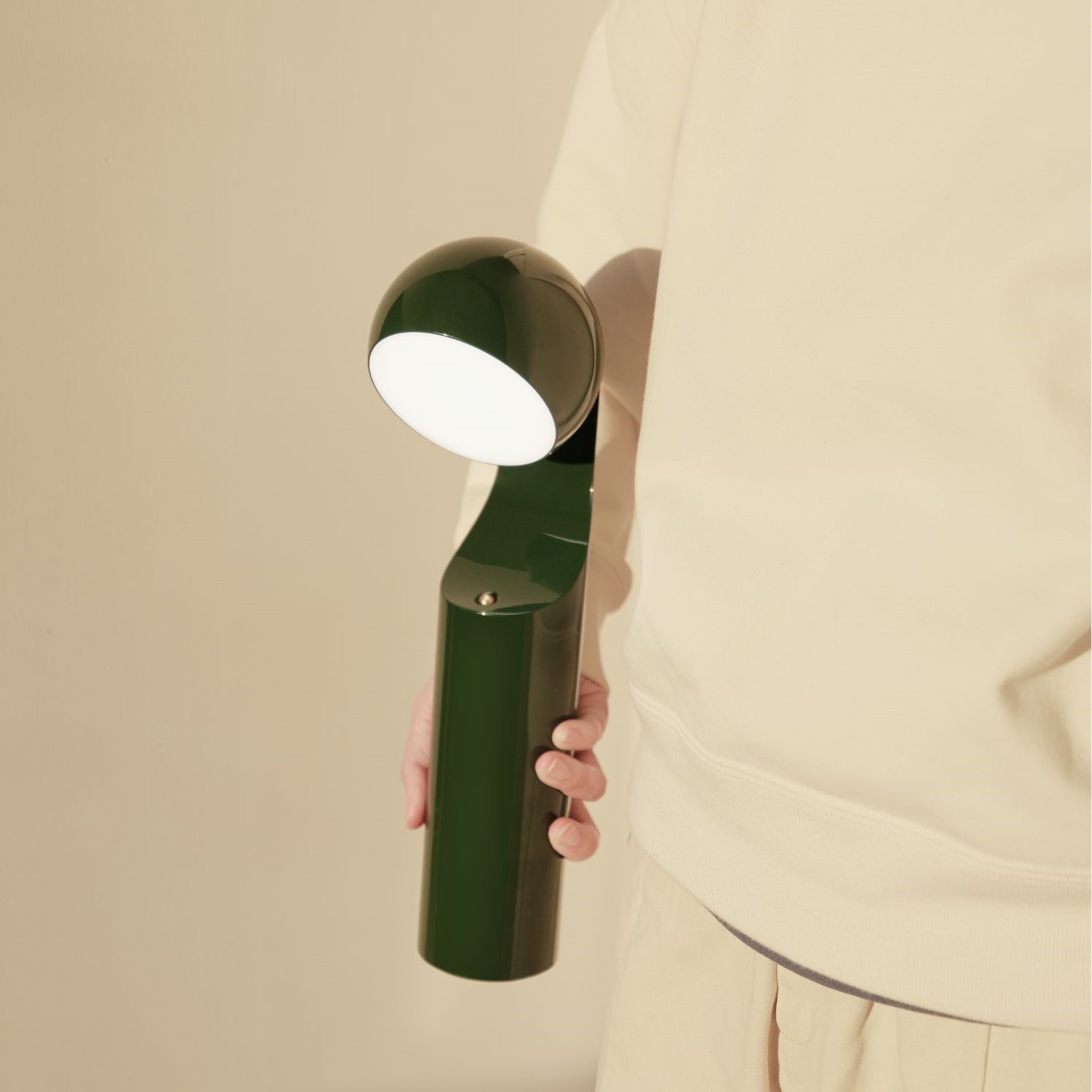 Een persoon in een lichtbeige outfit houdt de Fine Lumens Mono Leeslamp LED Oplaadbaar Forest Groen van Fine Lumens vast, met een ronde, verlichte kop, tegen een effen beige achtergrond.