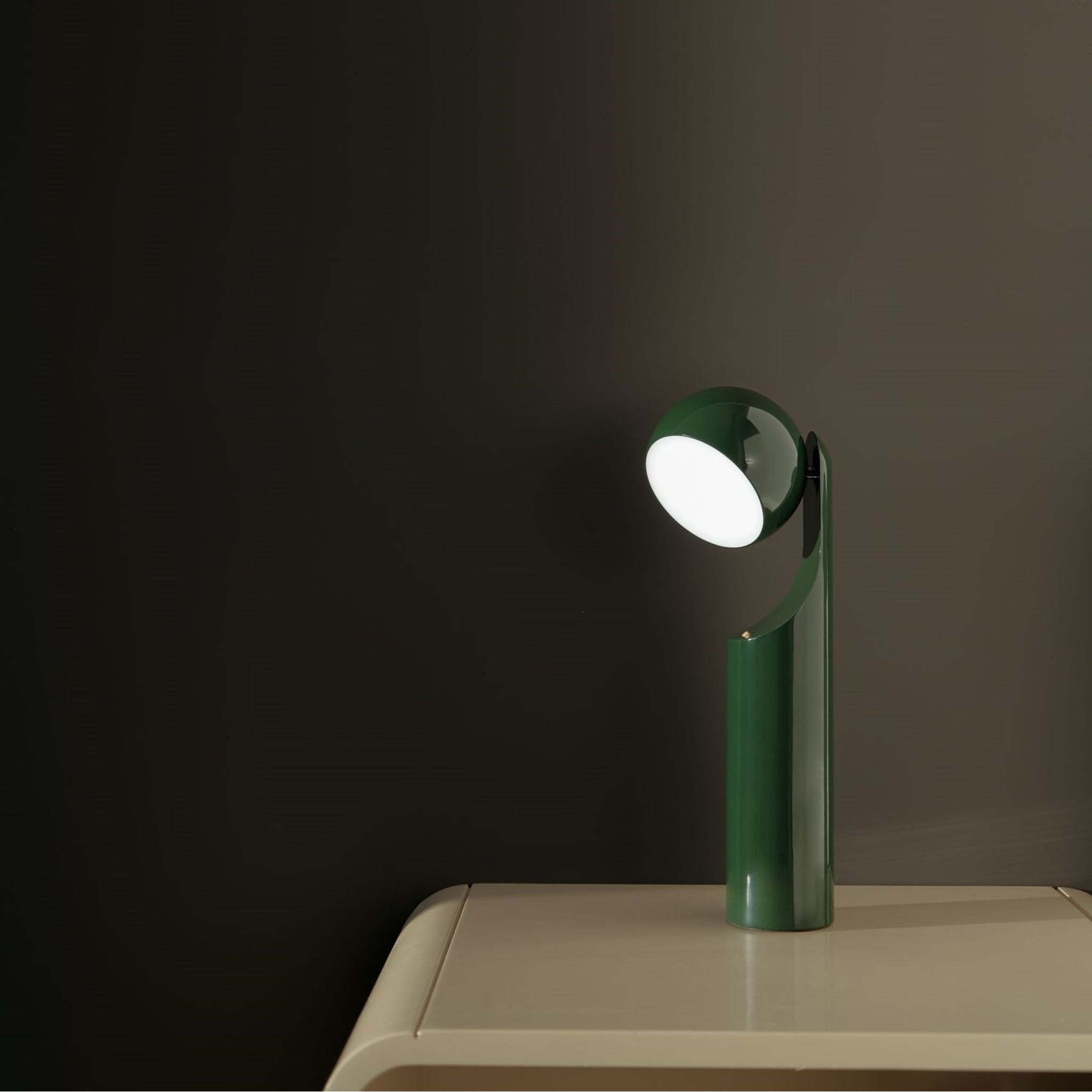 De Fine Lumens Mono Leeslamp LED Oplaadbaar in Forest Groen van Fine Lumens, met zijn moderne ronde kap, straalt op de hoek van een beige tafel tegen een donkere muur.