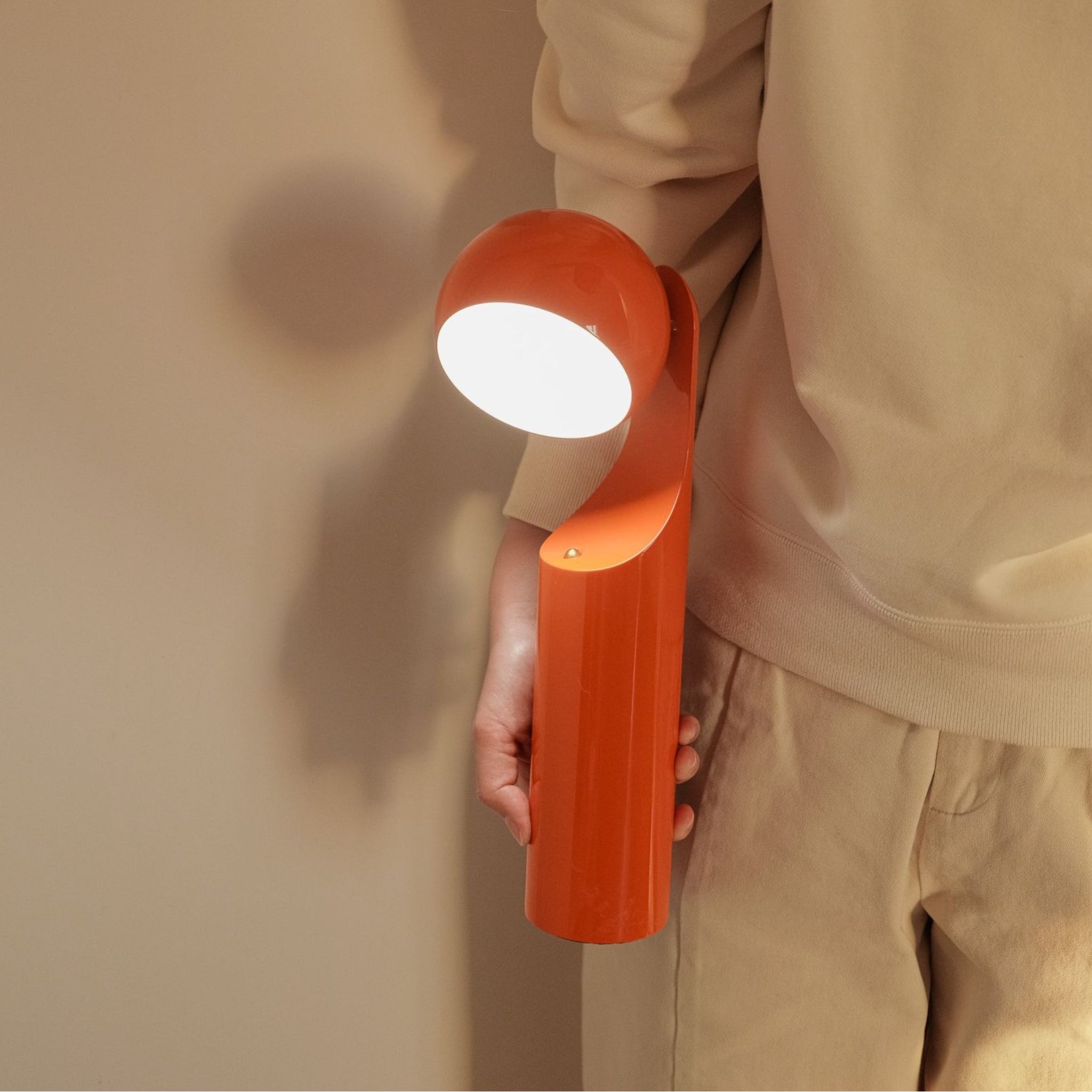 Een persoon in beige houdt de Fine Lumens Mono Leeslamp LED Oplaadbaar Mandarijn van Fine Lumens vast, een moderne en heldere oranje oplaadbare tafellamp met LED-verlichting, die een neutrale muur verlicht.
