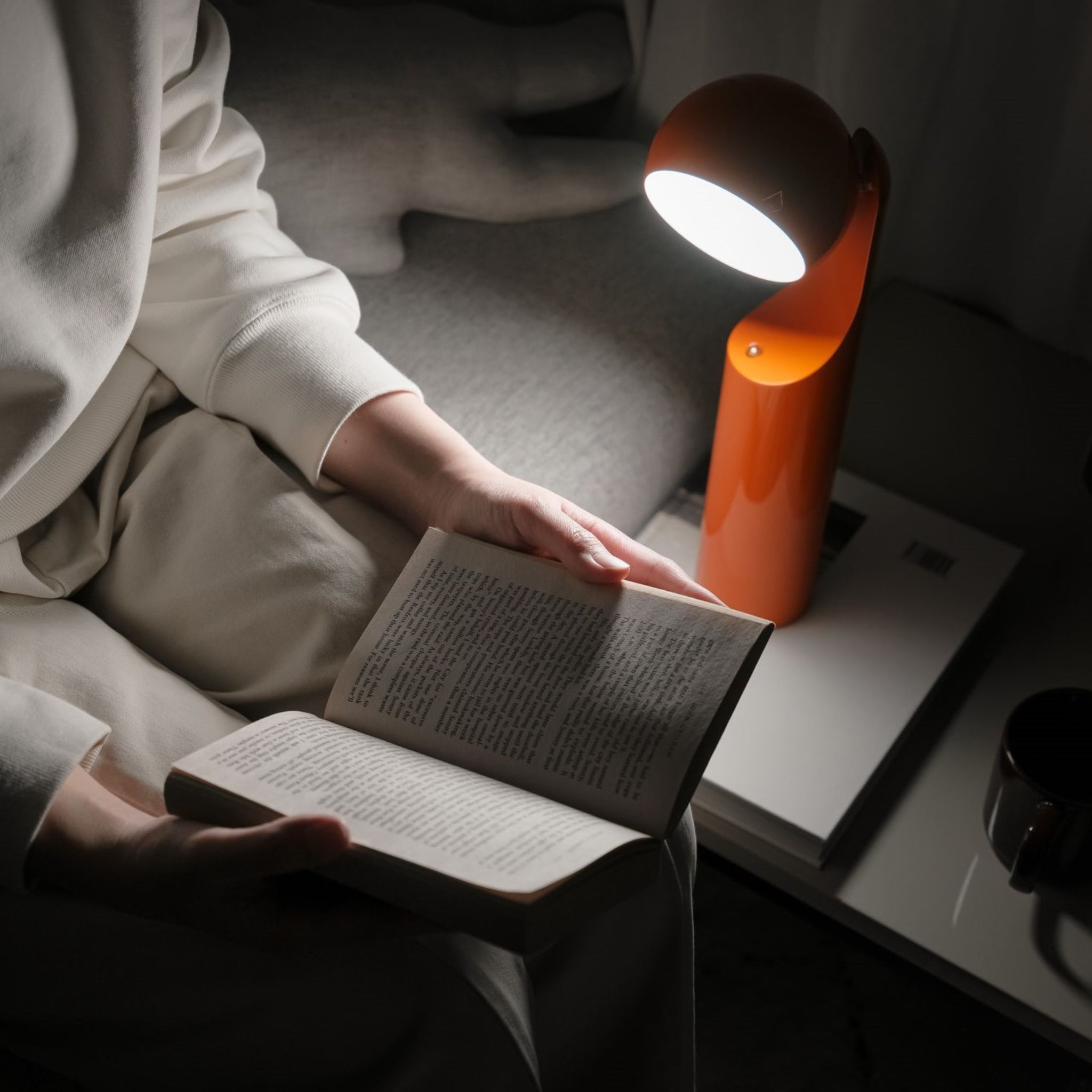 Een persoon in lichte kleding zit op een bank te lezen bij het schijnsel van de Fine Lumens Mono Leeslamp LED Oplaadbaar Mandarijn. Een kopje en een boek rusten vlakbij, waardoor er een rustige en gezellige sfeer ontstaat met deze moderne Fine Lumens lamp.