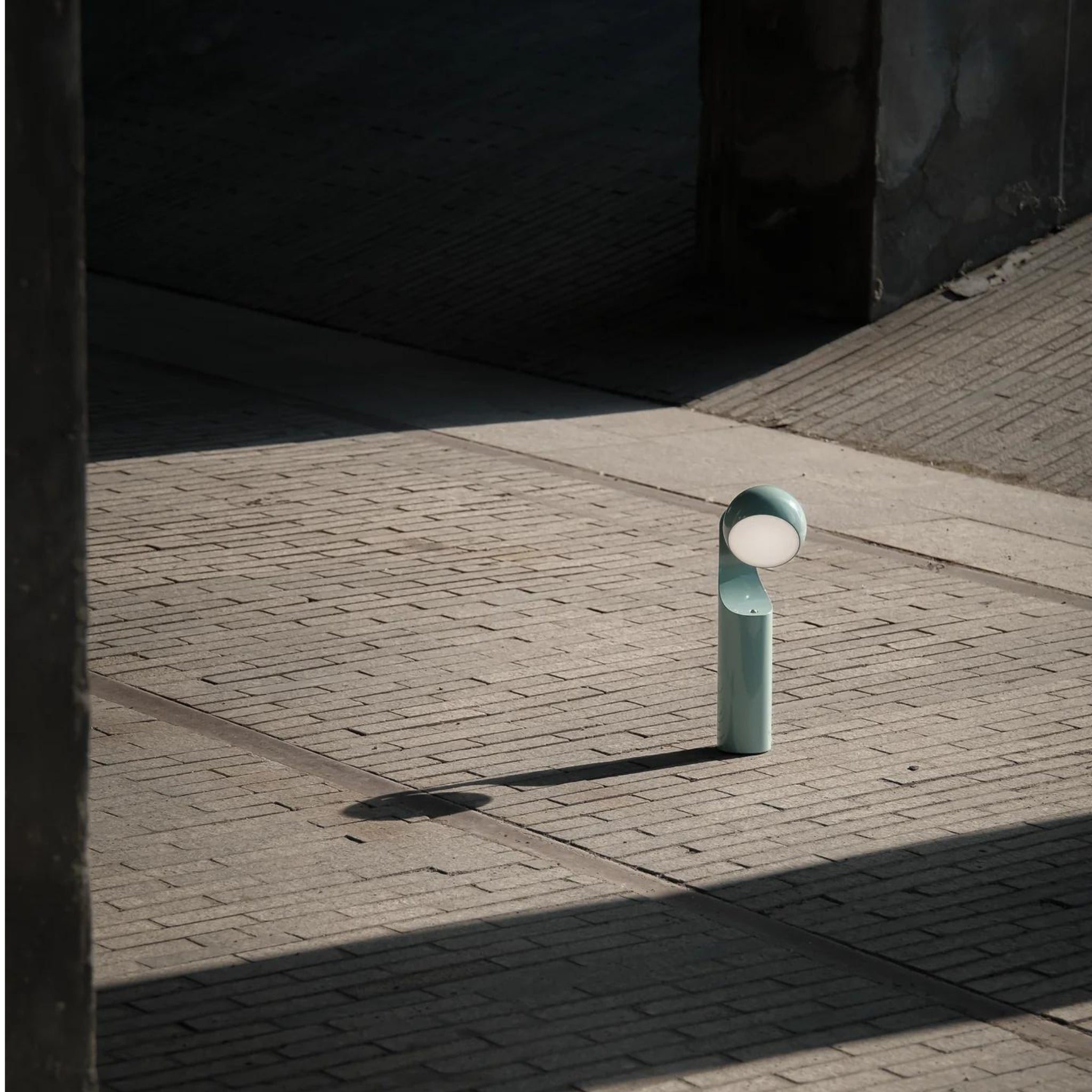 De Fine Lumens Mono Leeslamp LED Oplaadbaar Renew Blauw, een minimalistische tafellamp met een cilindrische voet en ronde bovenkant, werpt schaduwen op zonverlicht structuurbeton omgeven door geometrische vormen en donkere muren.