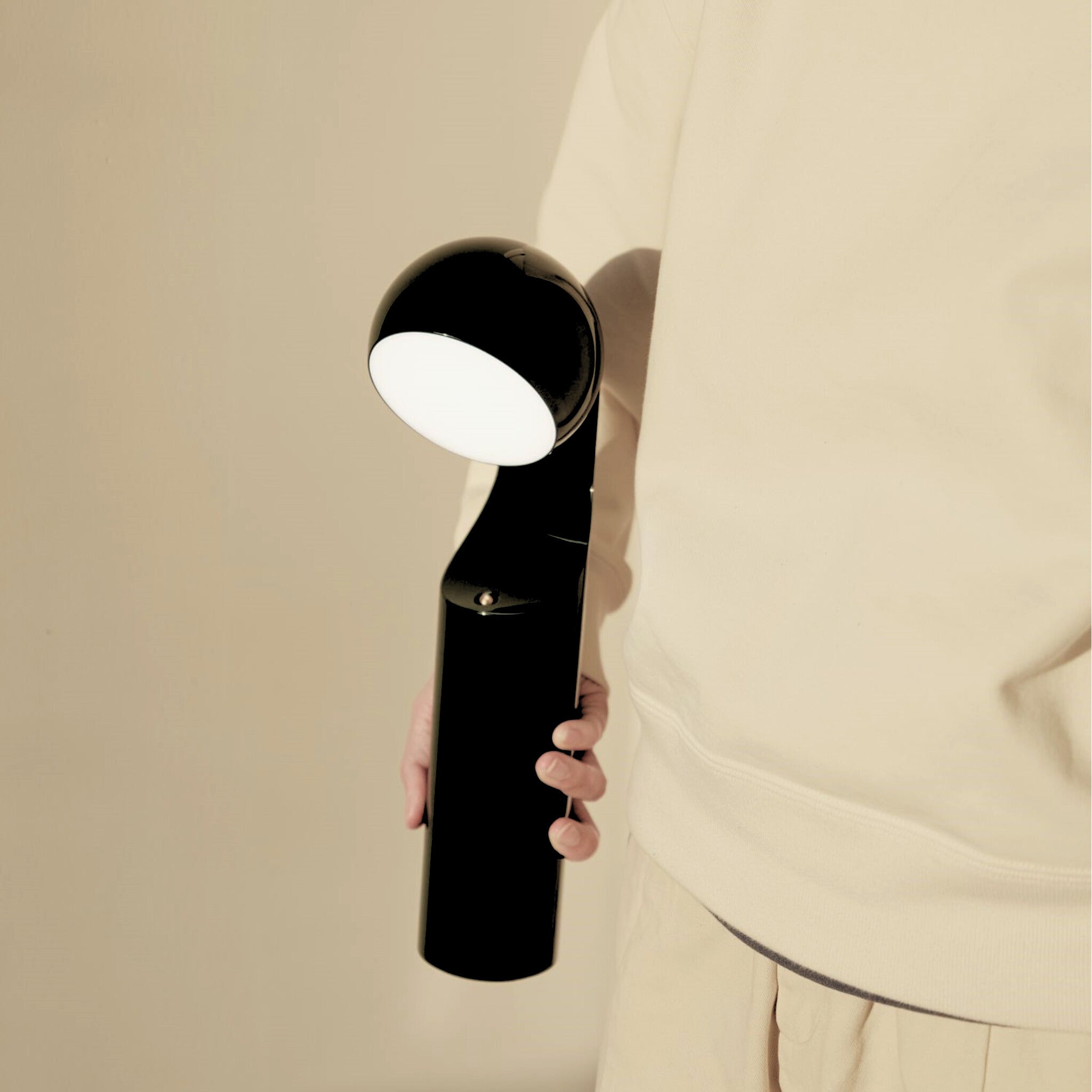 Een persoon in beige houdt de Fine Lumens Mono Leeslamp LED Oplaadbaar Renew Zwart vast, een moderne zwarte tafellamp van Fine Lumens met een rond gloeiend wit licht, tegen een neutrale achtergrond.