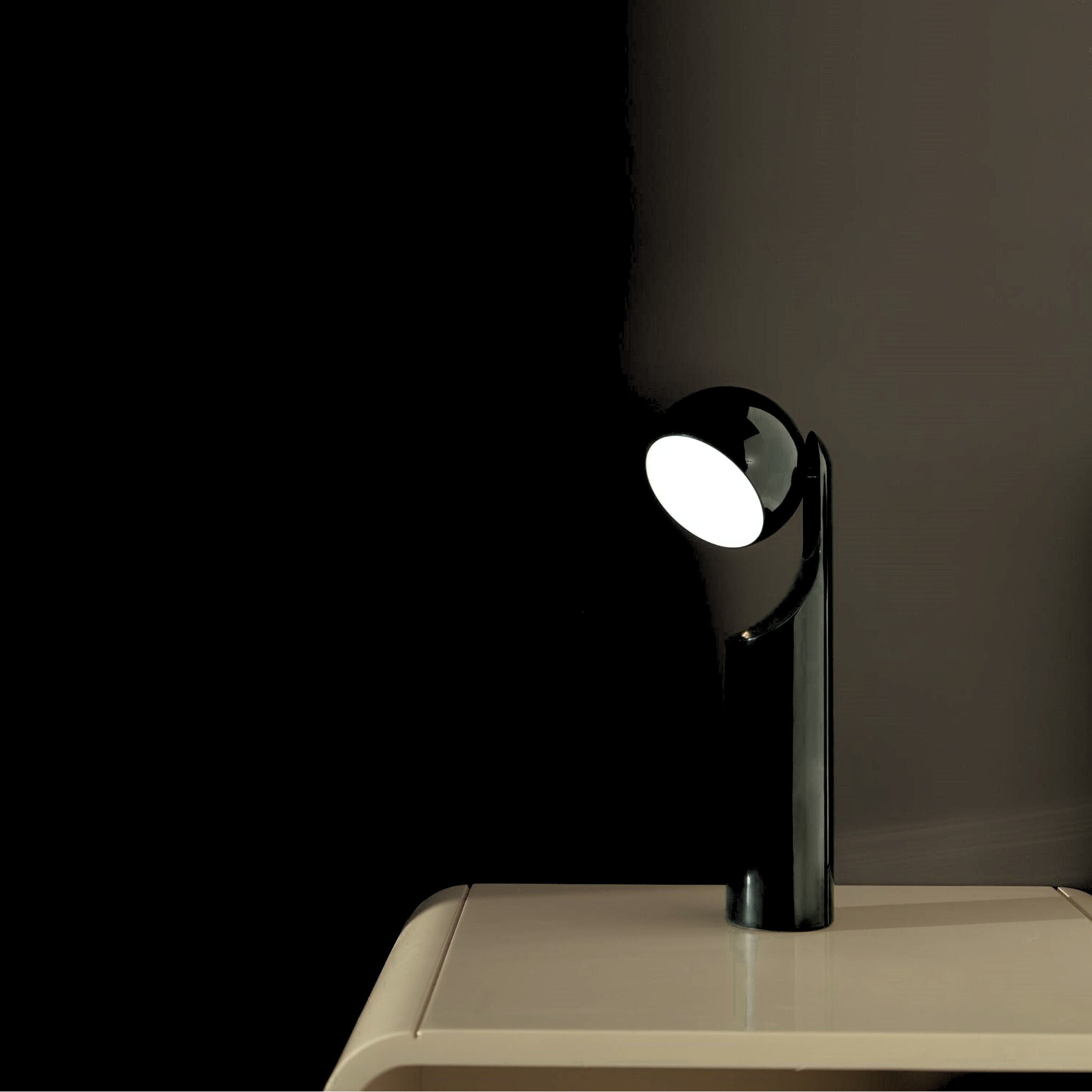 De Fine Lumens Mono Leeslamp LED Oplaadbaar Renew Zwart van Fine Lumens heeft een modern zwart ontwerp en een ronde kop, en werpt een zachte dimbare LED gloed op een beige tafel voor een rustige, eigentijdse look tegen een donkere achtergrond.