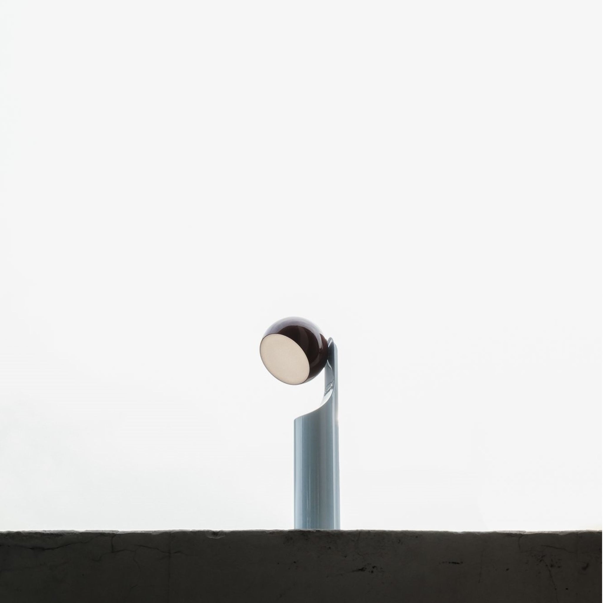 Een minimalistische foto van een enkele moderne straatlantaarn, een echo van de Fine Lumens Mono Leeslamp LED Oplaadbaar Roest Sky, die boven een betonnen barrière uitsteekt tegen een heldere, overbelichte witte lucht.