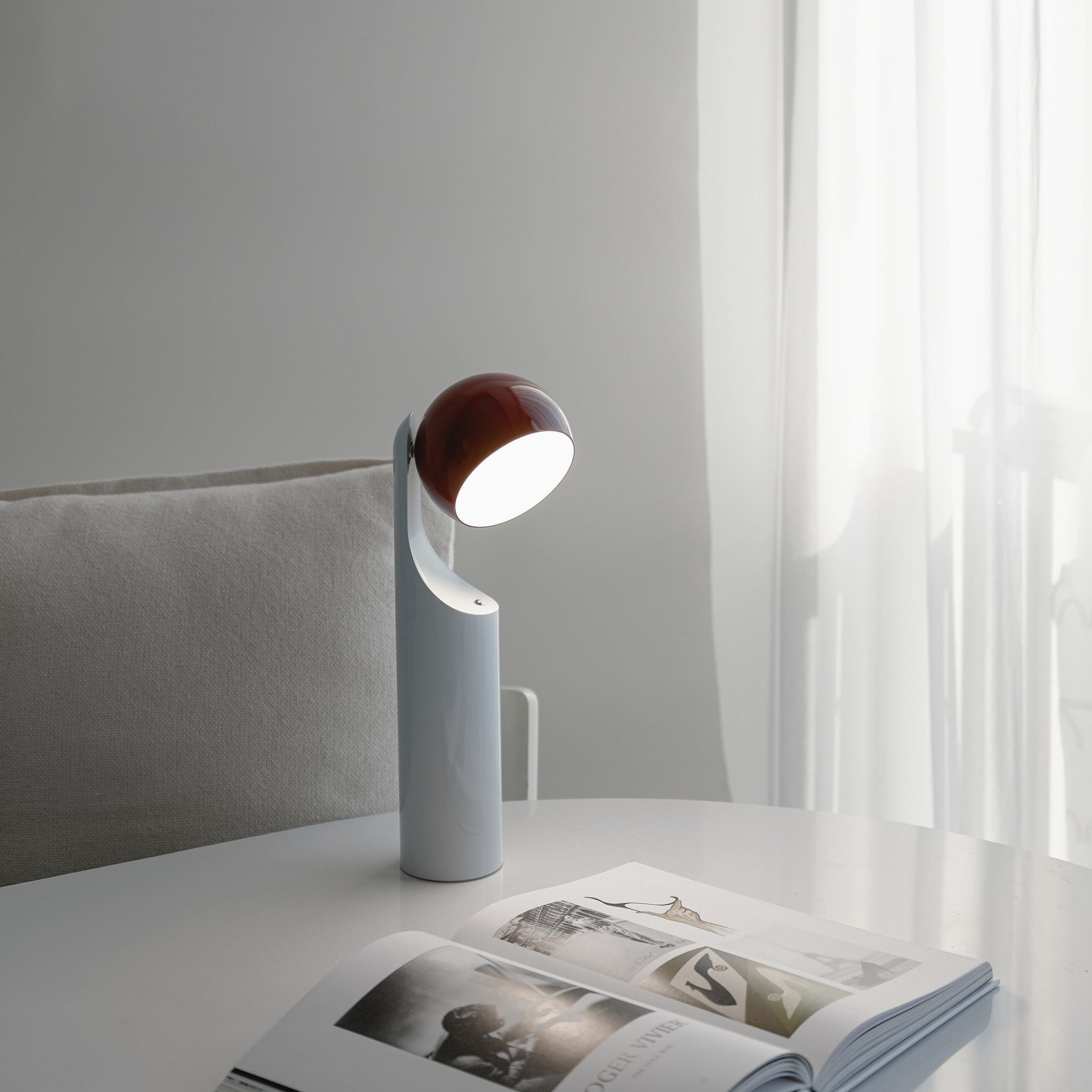 De Fine Lumens Mono Leeslamp LED Oplaadbaar Roest Sky van Fine Lumens werpt licht op een open tijdschrift op een witte tafel, met een lichtgrijze stoel met kussens en transparante gordijnen op de achtergrond, terwijl daglicht door het raam naar binnen valt.