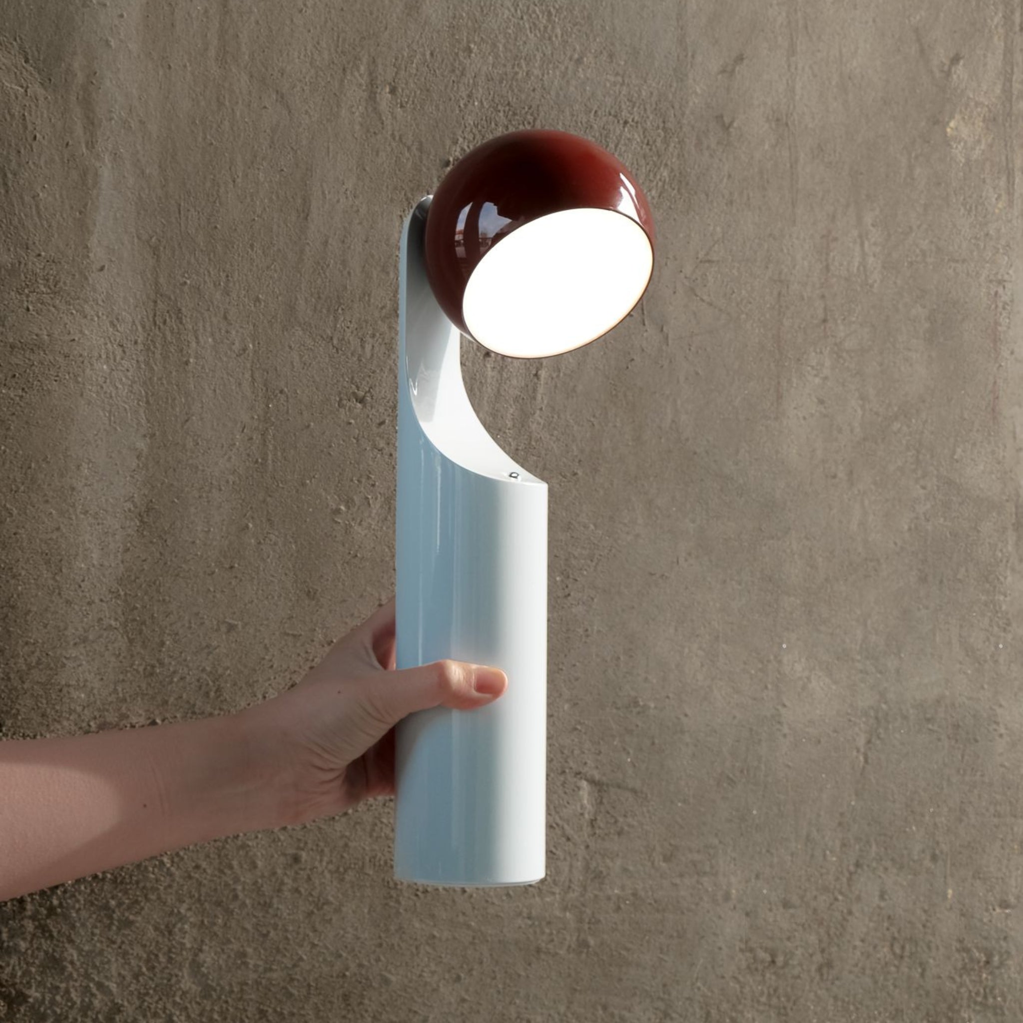 Een hand houdt een Fine Lumens Mono Leeslamp LED Oplaadbaar Roest Sky vast, met een ronde, verlichte kop in wit en rood, tegen een grijze muur met structuur.