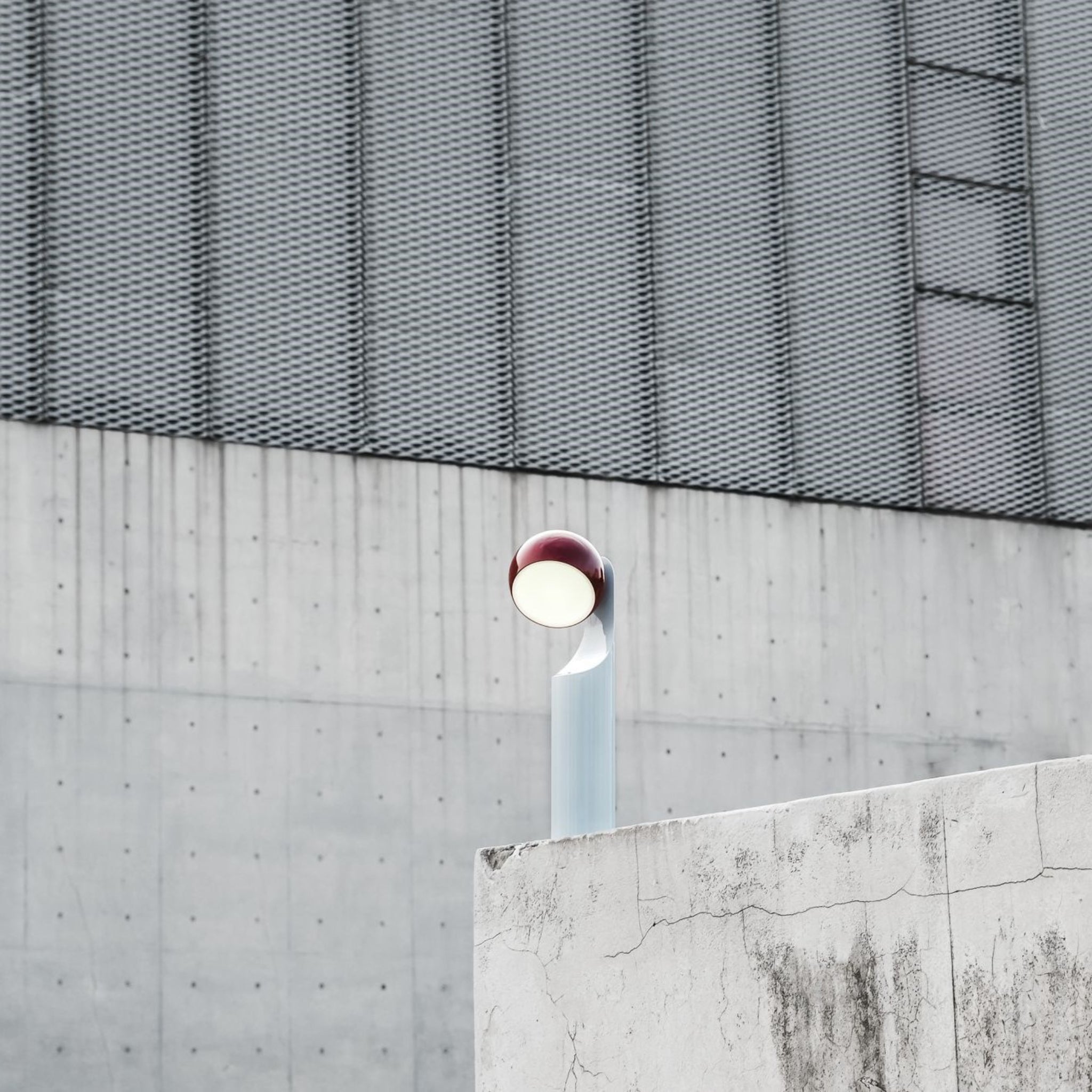 De Fine Lumens Mono Leeslamp LED Oplaadbaar Roest Sky van Fine Lumens, een moderne rood-witte dimbare LED-lamp, staat voor een groot grijs gebouw van beton en metaal met gebarsten, bevlekte muren op de voorgrond.