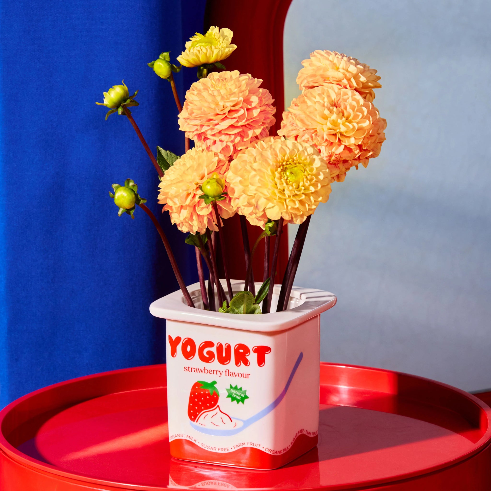 Een boeket lichtoranje bloemen in de Fluid Market Aardbei Yoghurt Vaas/Pot - een charmant stuk van keramiek - rust op een rode tafel met blauwe en witte stof, wat een speels accent toevoegt aan elke Fluid Market vitrine.